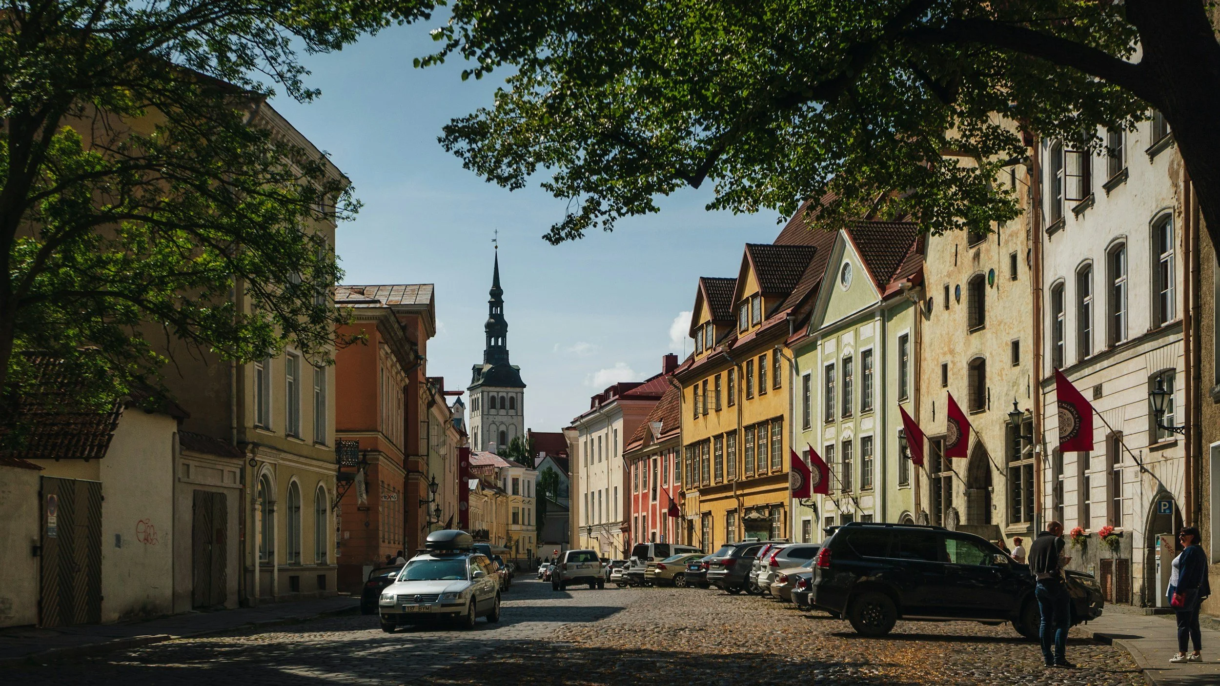 A European city street with colorful historic buildings, parked cars, and two pedestrians, with a church spire in the background and trees lining the street.