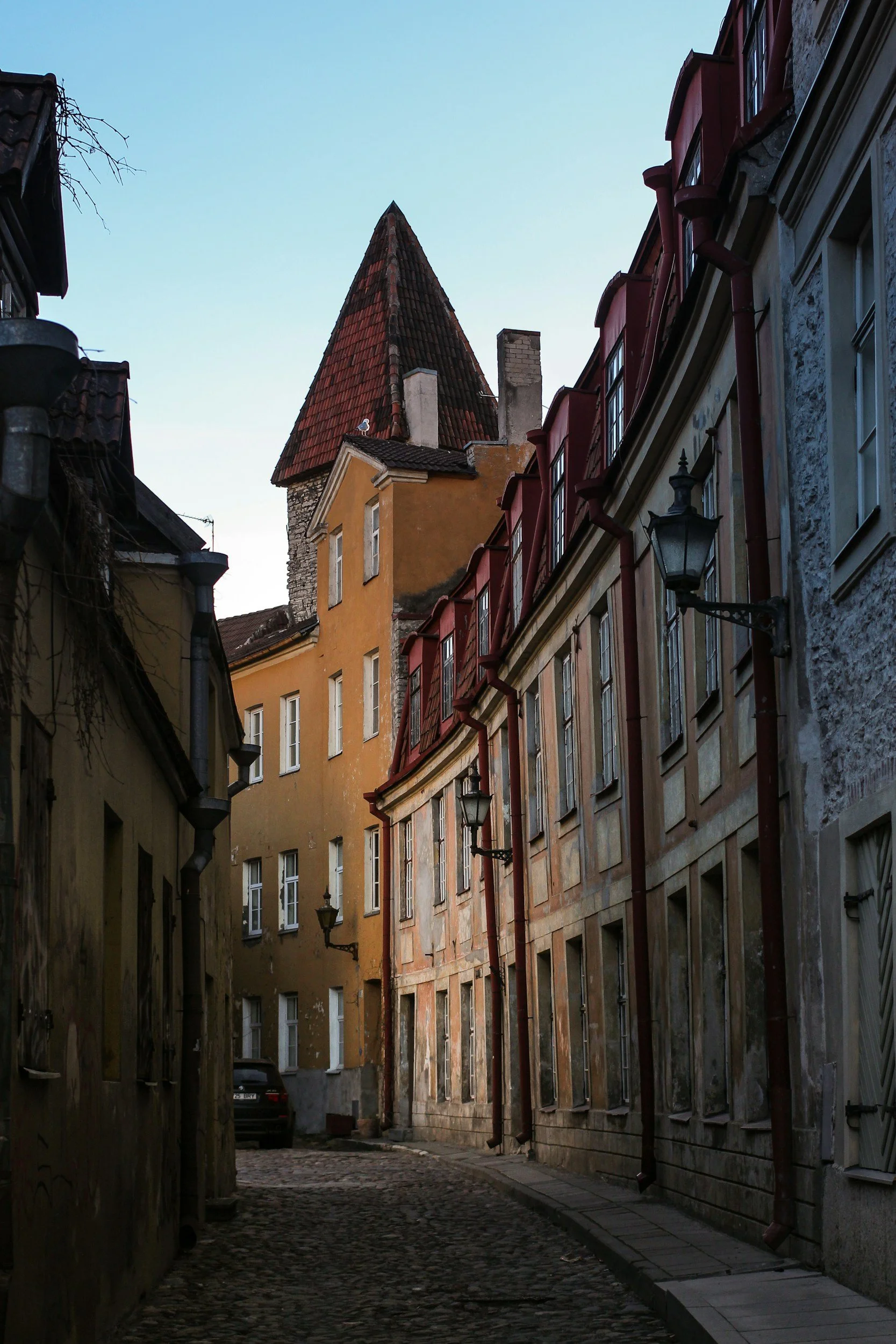 A narrow cobblestone street lined with old buildings with colorful facades, and a tall, steep, red-tiled roof tower in the background against a pale blue sky.