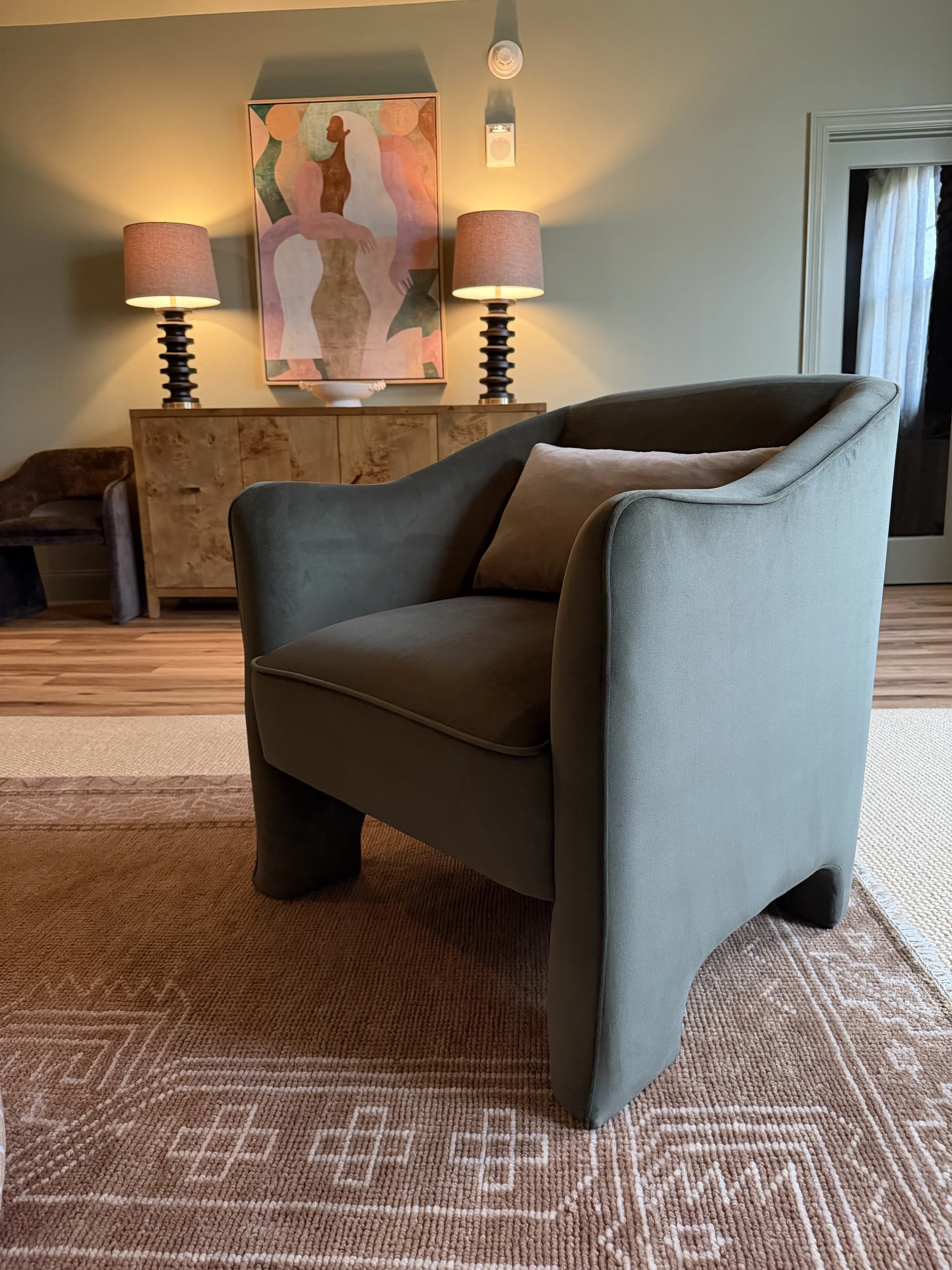 Green velvet armchair on a patterned rug, with lamps and art behind it