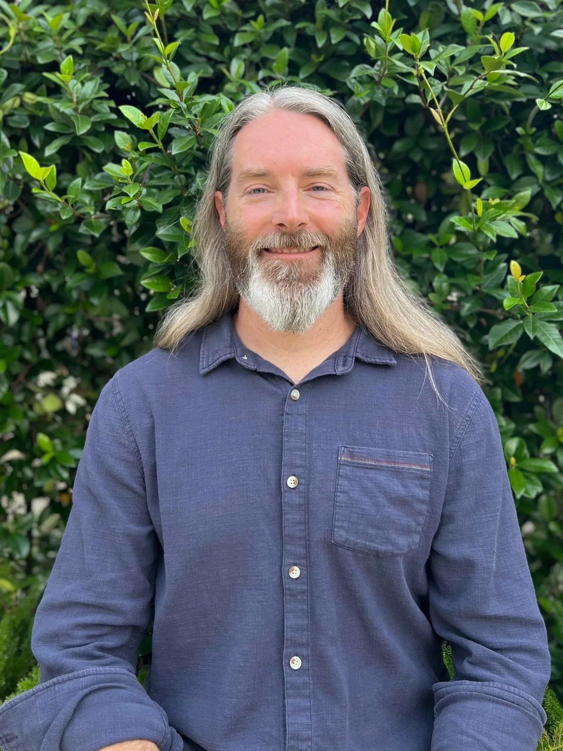 A smiling man with long gray hair and a beard, wearing a dark blue button-up shirt, standing outdoors in front of a leafy green hedge.