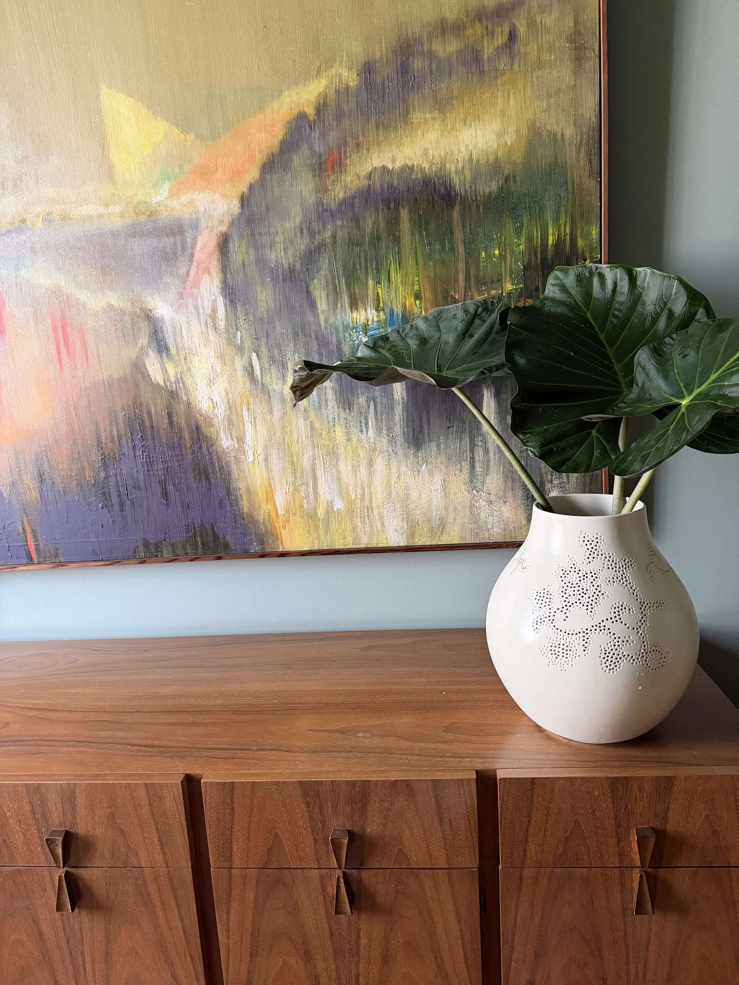 Walnut dresser with a white vase of large leaves beneath an abstract landscape painting