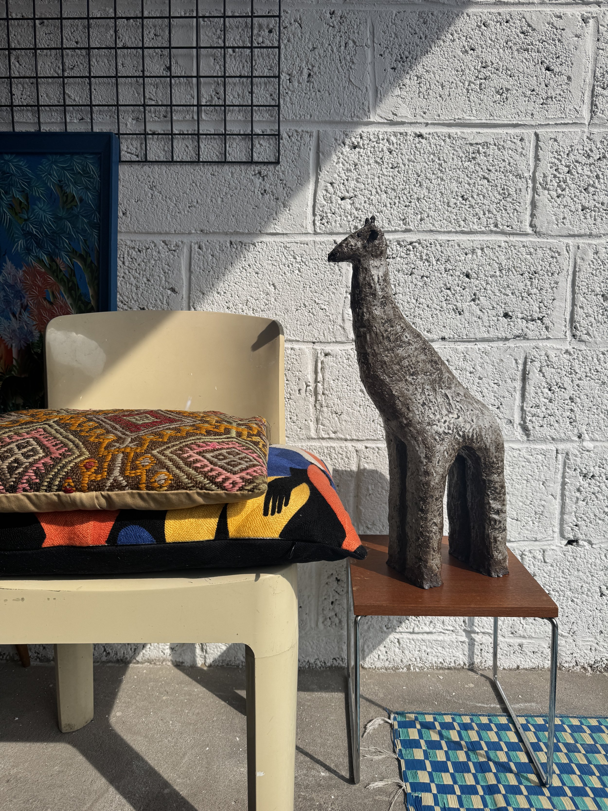 Decorative scene with a cream-colored plastic chair, colorful cushions, a small wooden table with a textured animal sculpture, a multicolored woven rug, and a white brick wall with a grid and artwork.