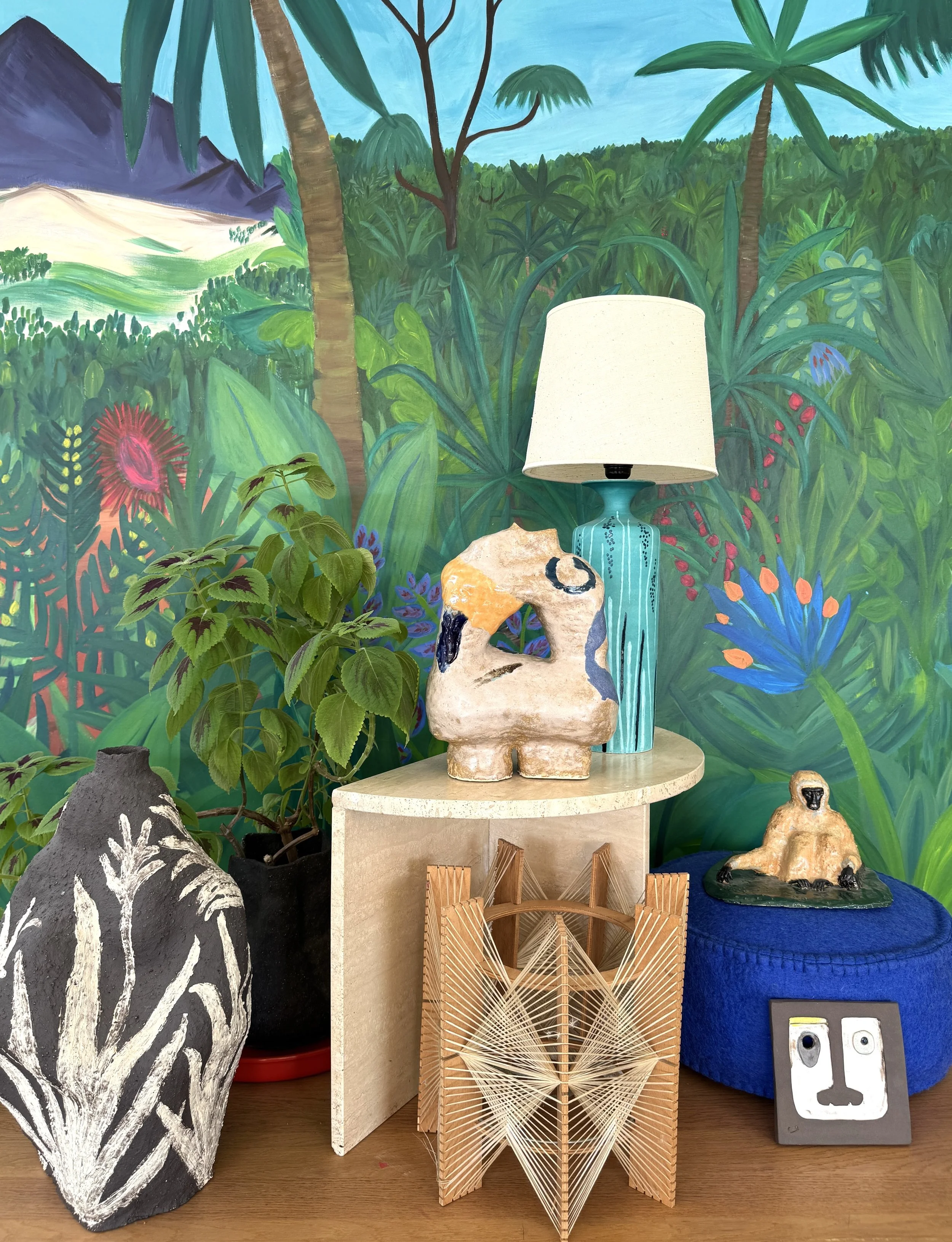 Decorative items on a wooden table in front of a vibrant jungle-themed mural. Items include a black and white painted vase, a potted plant with green leaves, a abstract ceramic sculpture, a blue table lamp with a white shade, a wooden geometric art piece, a small seated figurine, and a colorful artwork of a face.