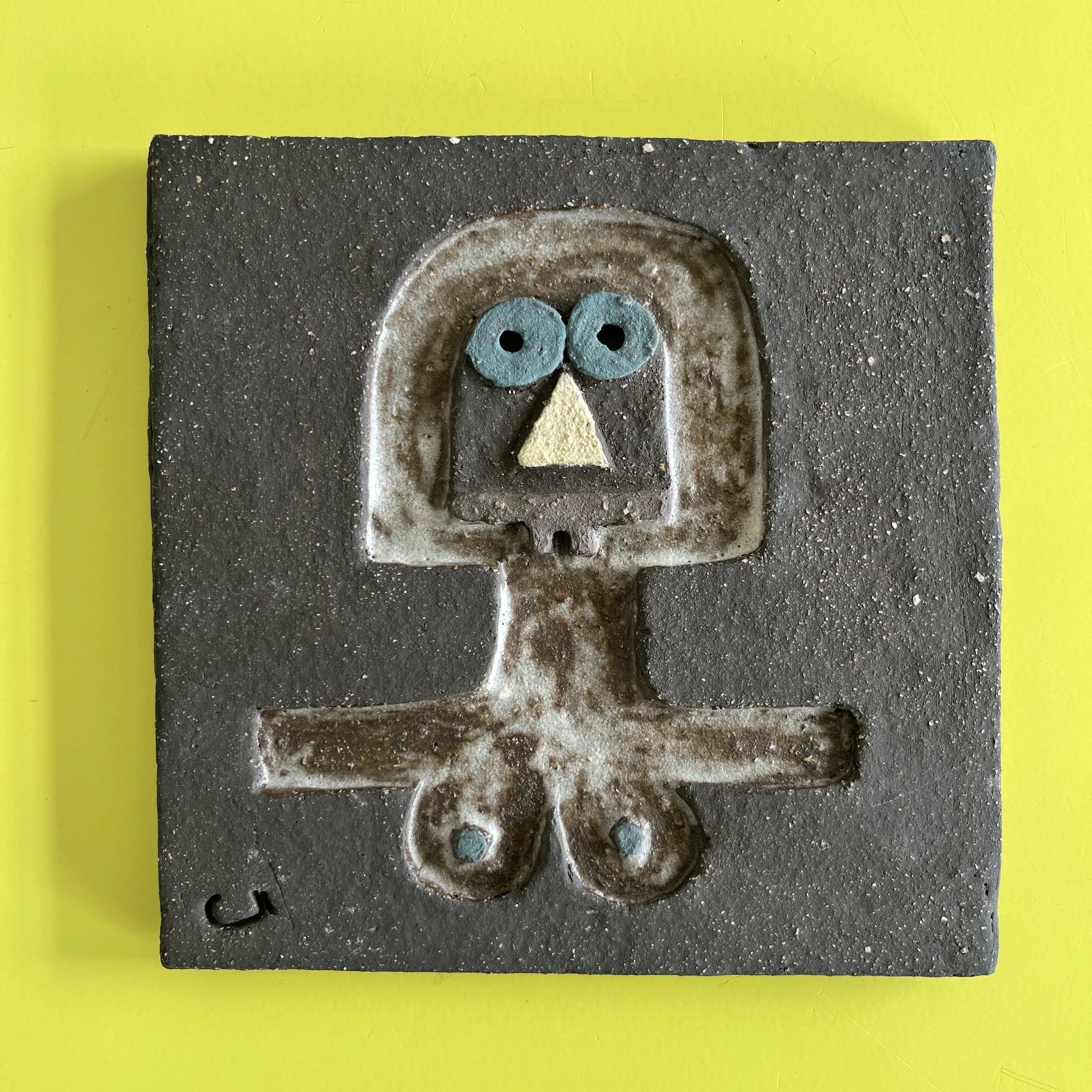 Decorative ceramic wall art with abstract face design, featuring yellow and pink eye shapes and a small smiling mouth on a black background.