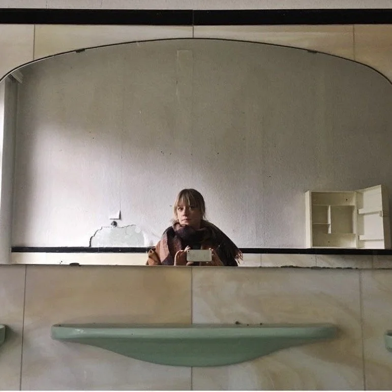 A woman taking a mirror selfie in an abandoned or rundown bathroom with cracked and peeling walls, and empty shelves in the background.