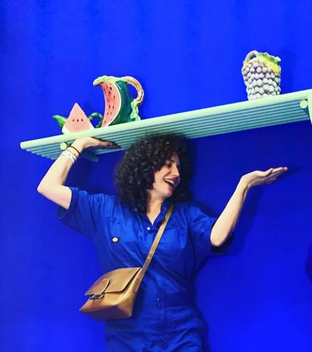 Woman posing with a shelf on her shoulder holding colorful, sculpted fruit.