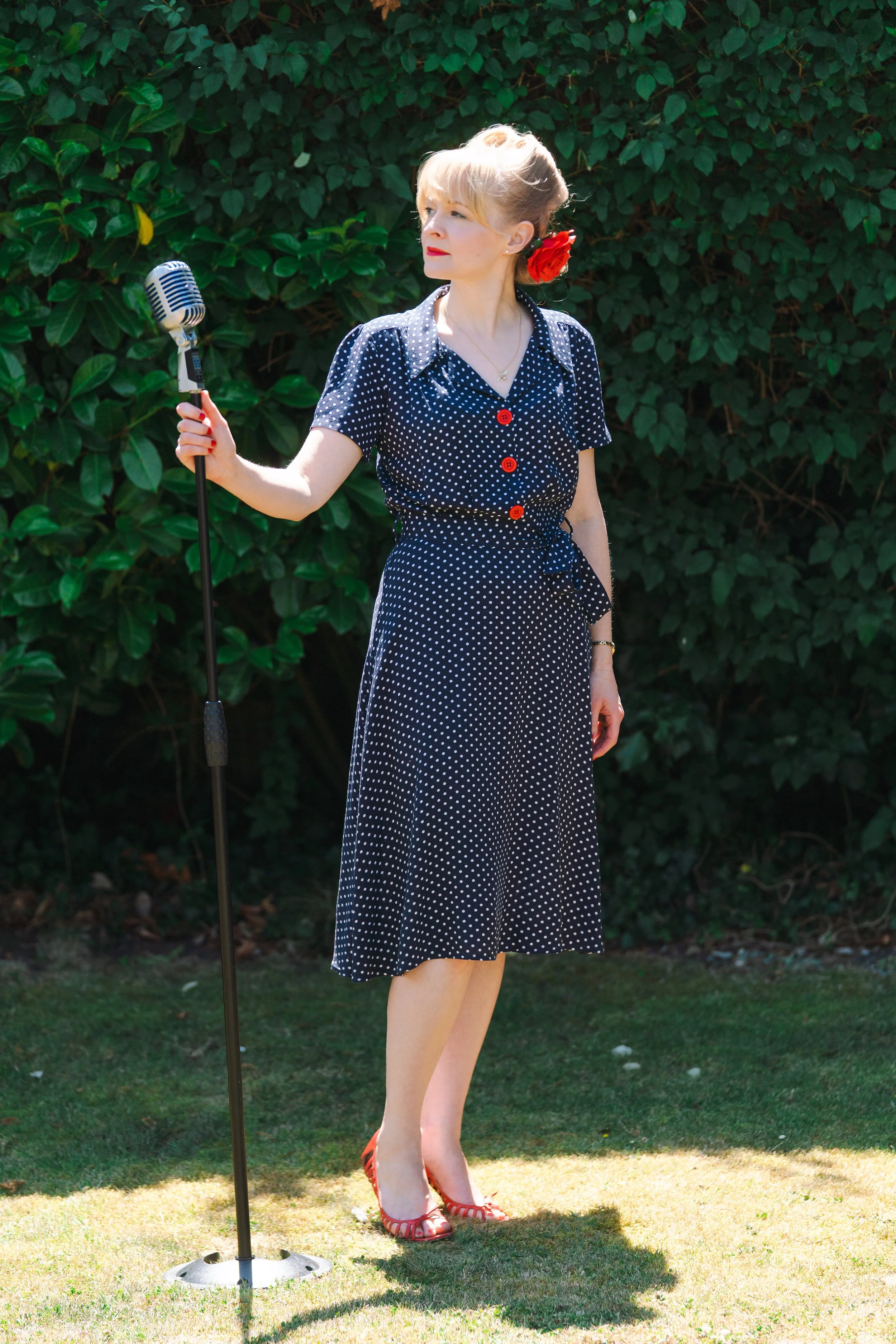 1940s wartime singer Kitty Mazinsky stands on a lawn wearing a 1940s-style shirt-dress in navy with white polka dots and red buttons. Her hair is swept up in victory rolls with a red rose pinned behind one ear, and she is holding a vintage-style micr