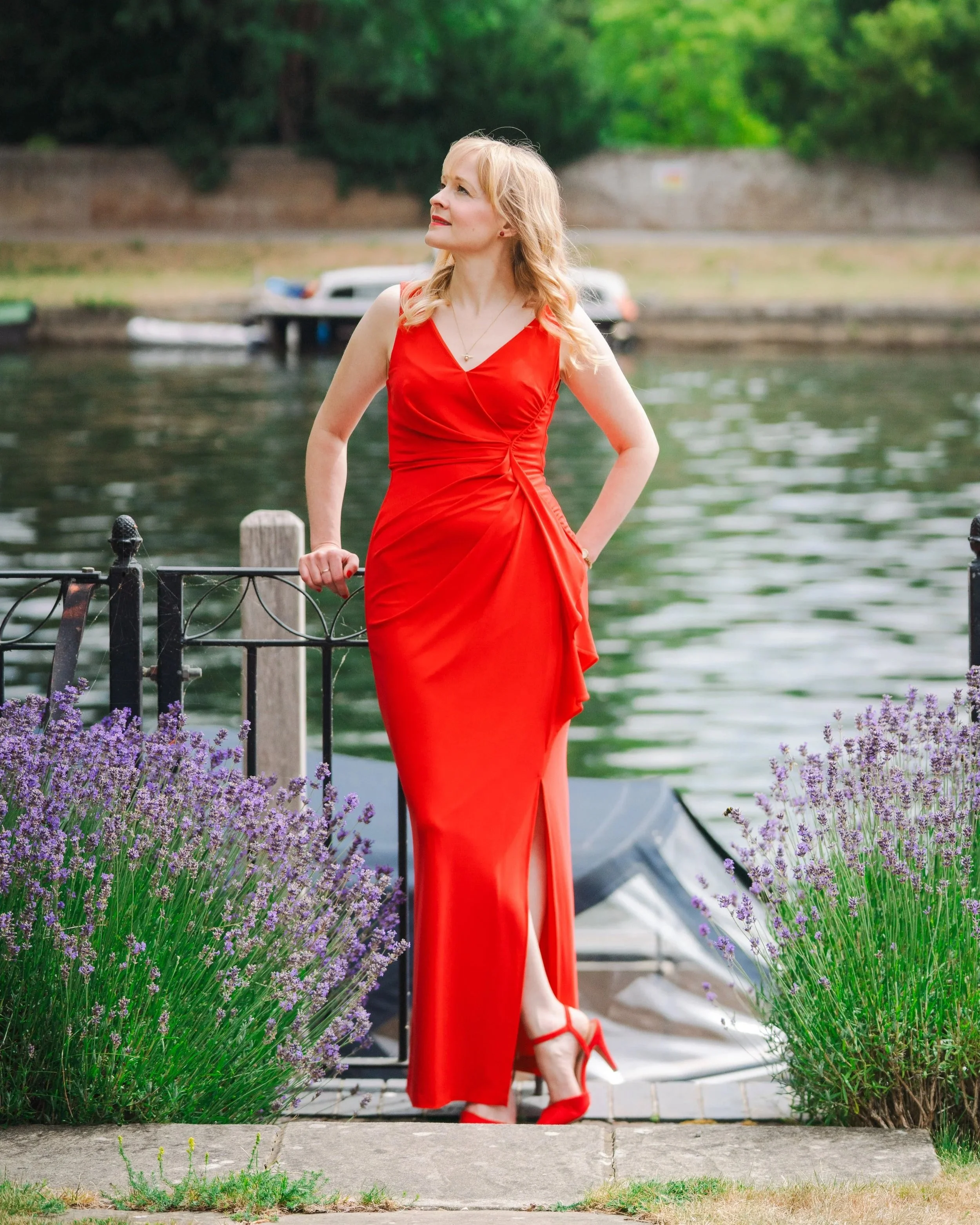 Kitty Mazinsky leans on a metal railing between lavender bushes in front of a boat, wearing a red full-length dress, with a river flowing behind her.