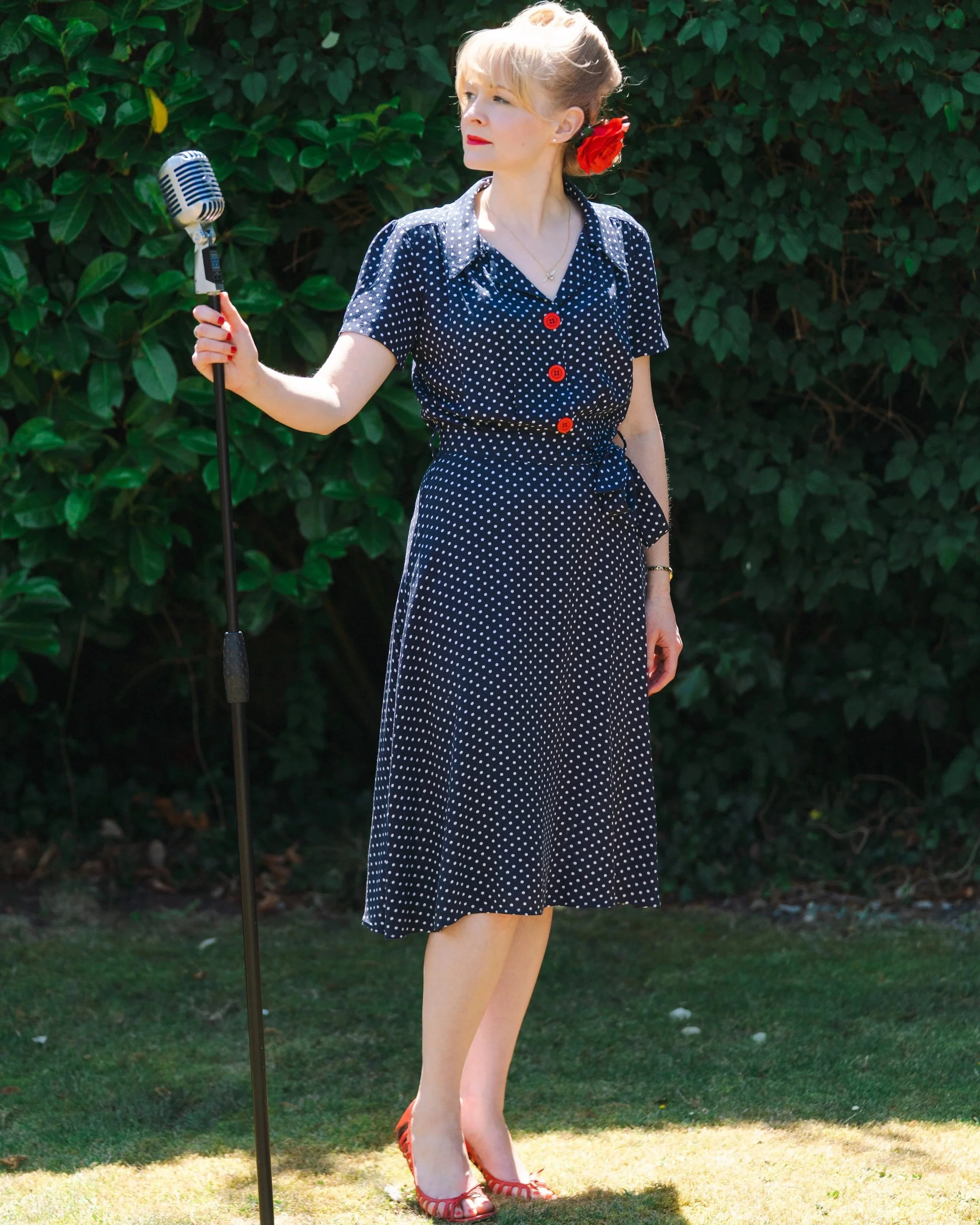 Vintage singer Kitty Mazinsky stands on a lawn in front of a tall bush, wearing a 1940s-style shirt-dress in navy with white polka dots and red buttons. Her hair is swept up in victory rolls with a red rose pinned to one side, and she is holding a Sh
