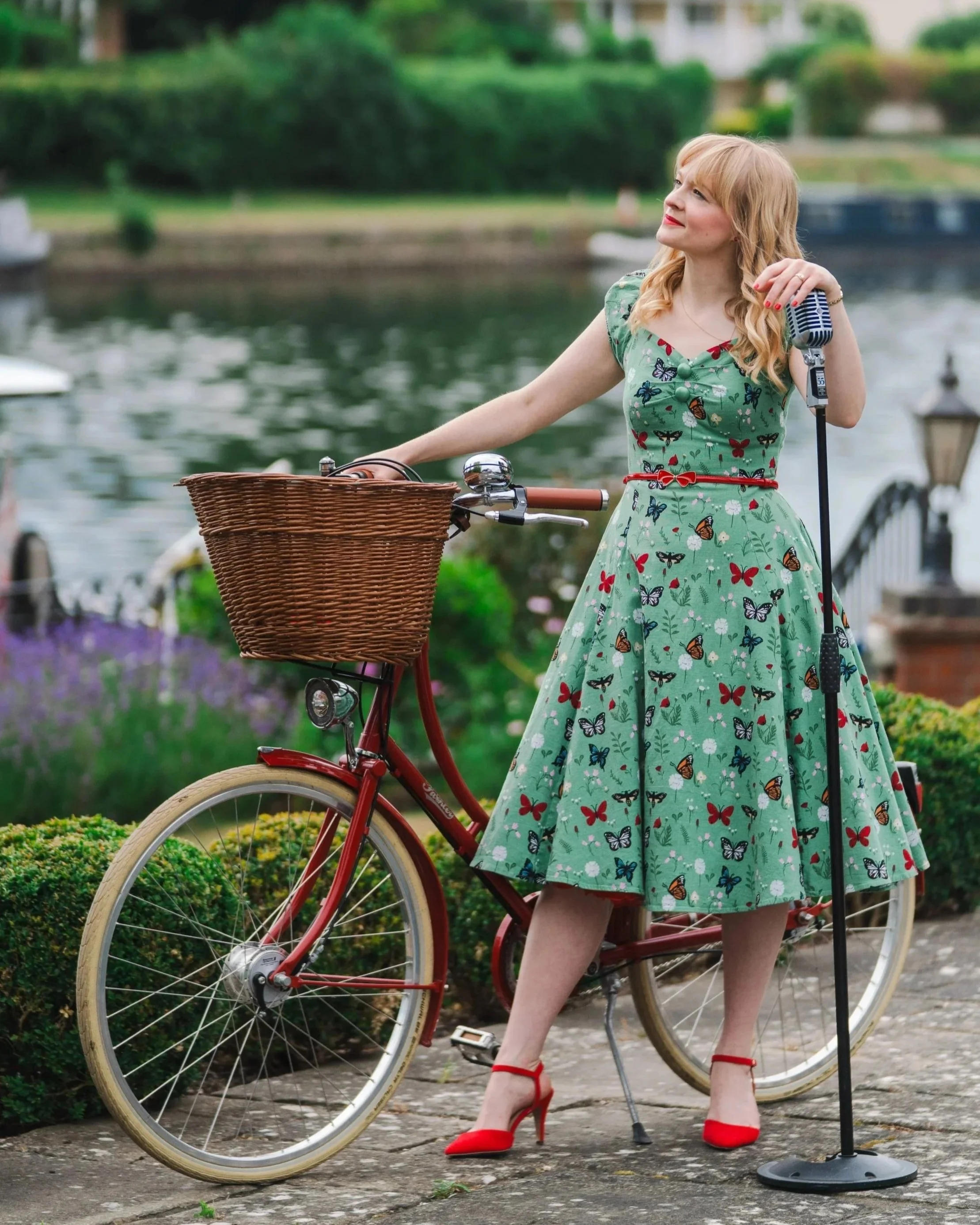 Kitty Mazinsky leans on a Shure 55 vintage-style microphone, wearing a green butterlfy patterned 1950s-style dress with puffed sleeves, a cinched waist and a tea-dress-style skirt. She is holding a red Pashley cycle with a wicker basket. Behind her a