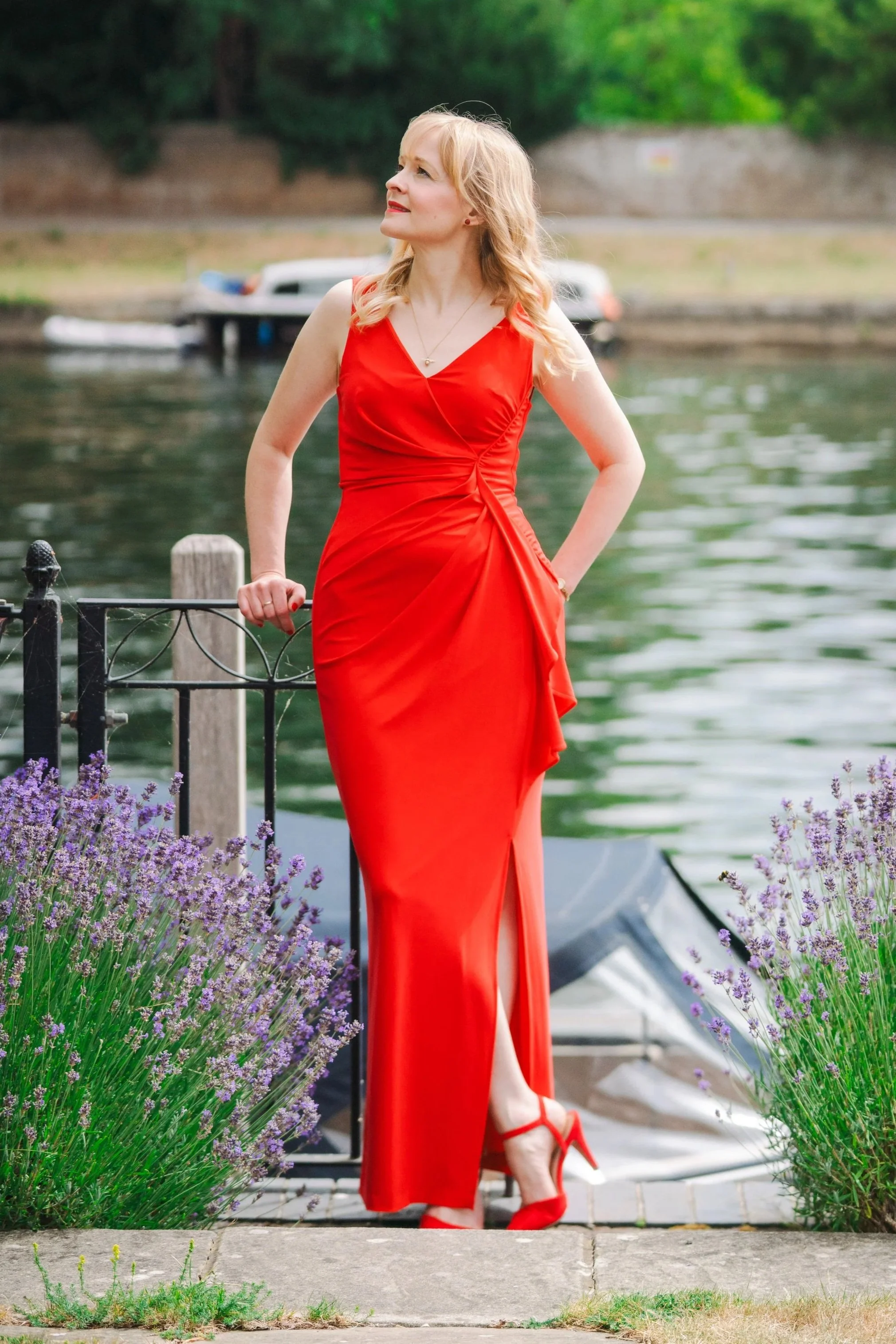 Kitty Mazinsky leans on a metal railing between two lavender bushes on the bank of the river Thames, with two boats in the background. She wears a red full-length dress, with the river flowing behind her.