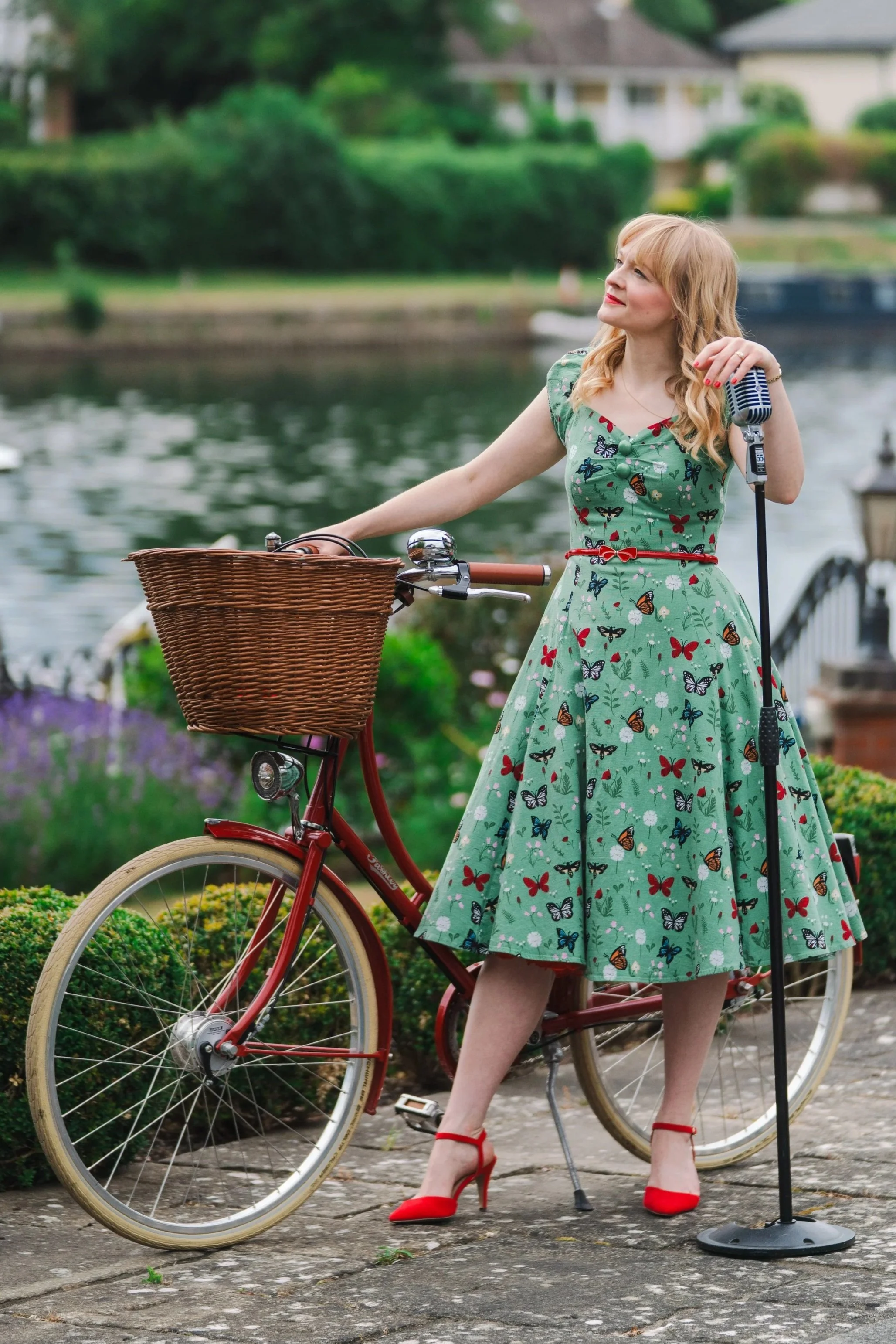 Vintage jazz vocalist Kitty Mazinsky leans on a Shure 55 vintage-style microphone, wearing a green butterfly-patterned 1950s-style dress with puffed sleeves, a cinched waist and a tea-dress-style skirt. She is holding a red Pashley bicycle with a wic