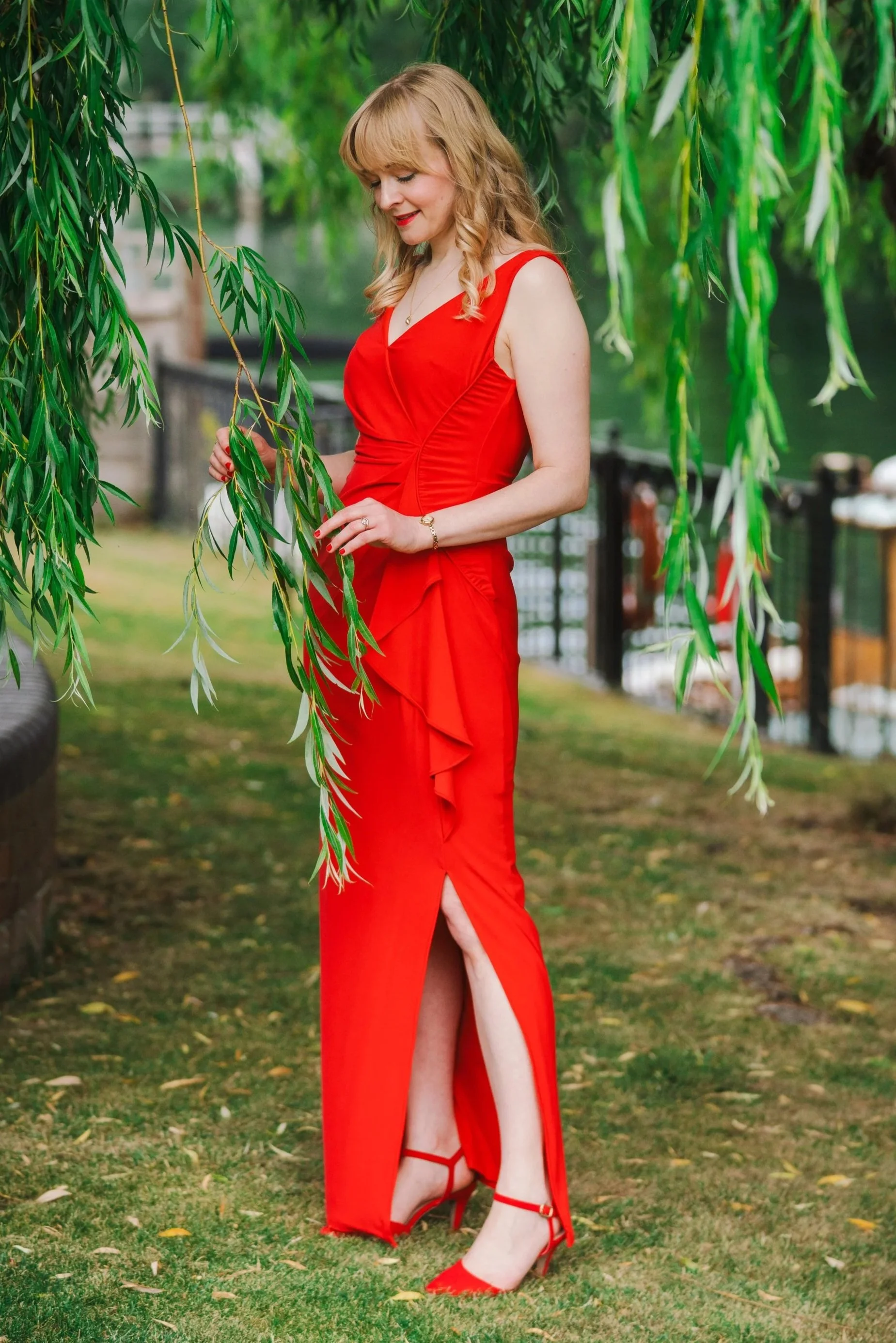 Kitty Mazinsky stands on a lawn among the fronds of a weeping willow tree, holding the leaves between her forefingers. In the background is a 1925 wooden pleasure craft moored on the banks of the river Thames. She wears a red, floor-length dress.