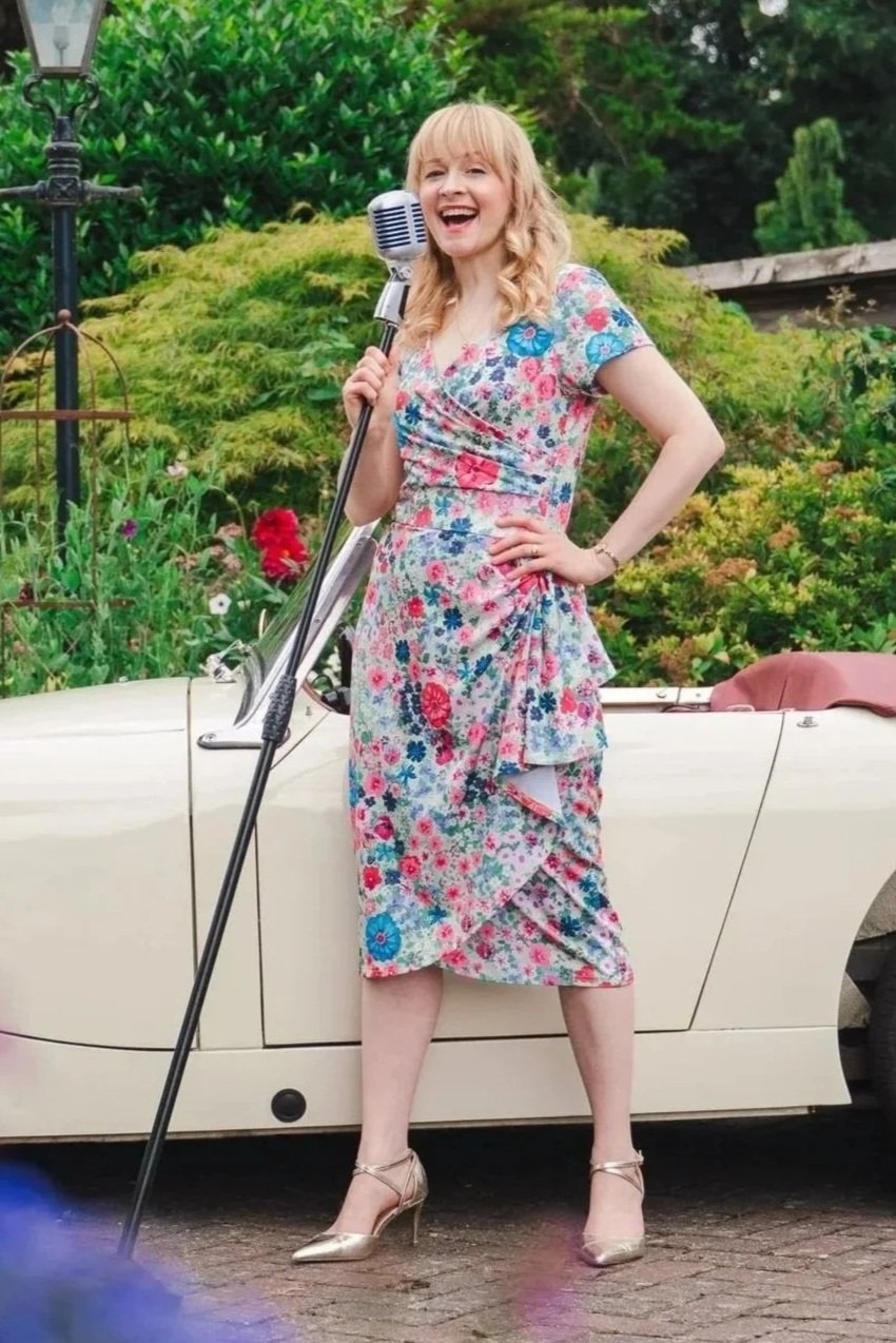 Vintage jazz vocalist Kitty Mazinsky holds a Shure 55 vintage-style microphone on a mic stand, leaning on the side of a cream Frogeye Sprite parked on a red-brick drive. In the background are trees and an old-fashioned lamppost. She wears a floral dr