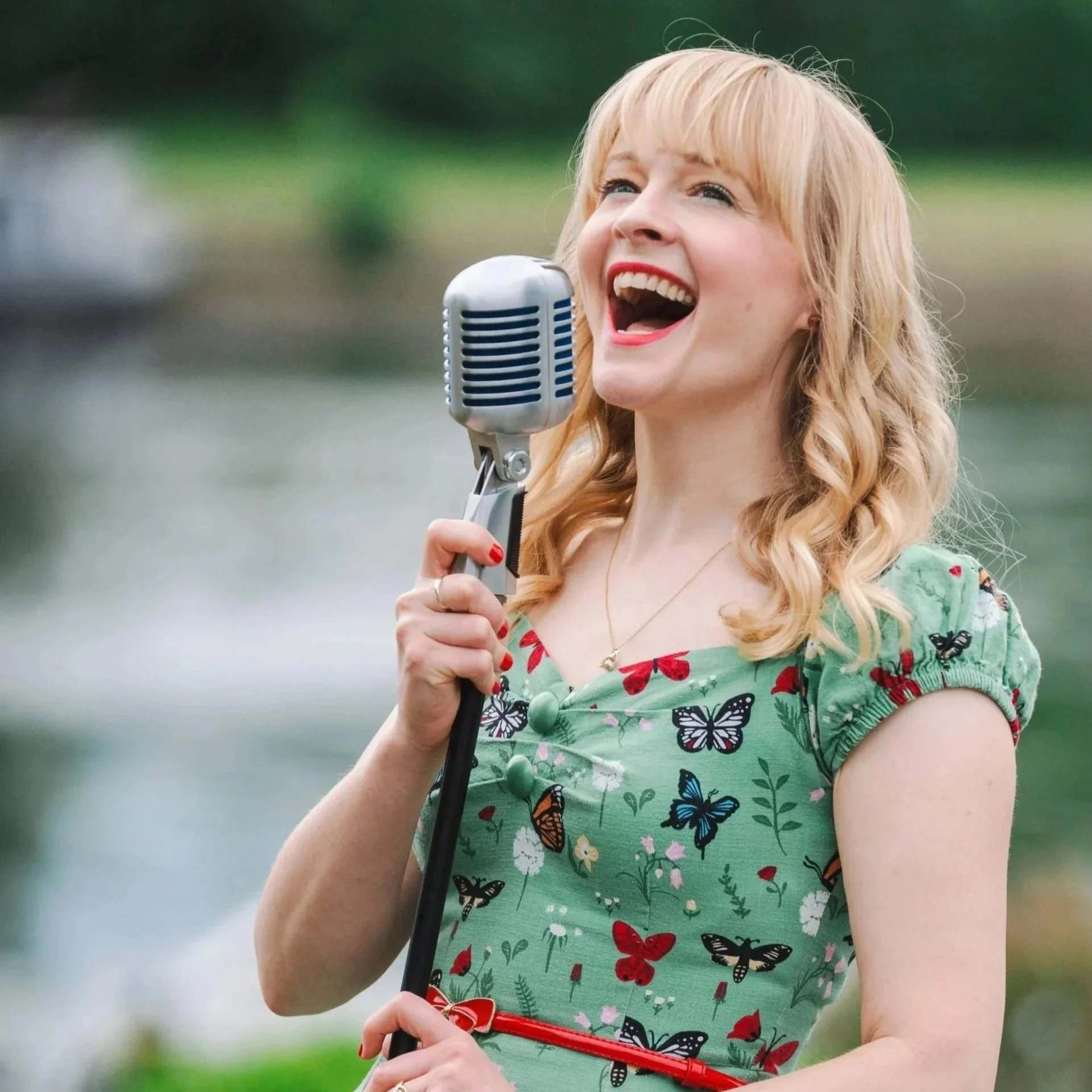 Kitty Mazinsky sings into a vintage-style Shure 55 microphone. She has a broad smile and is wearing a green dress with cap sleeves and a 1950s silhouette, featuring a butterfly and floral motif.