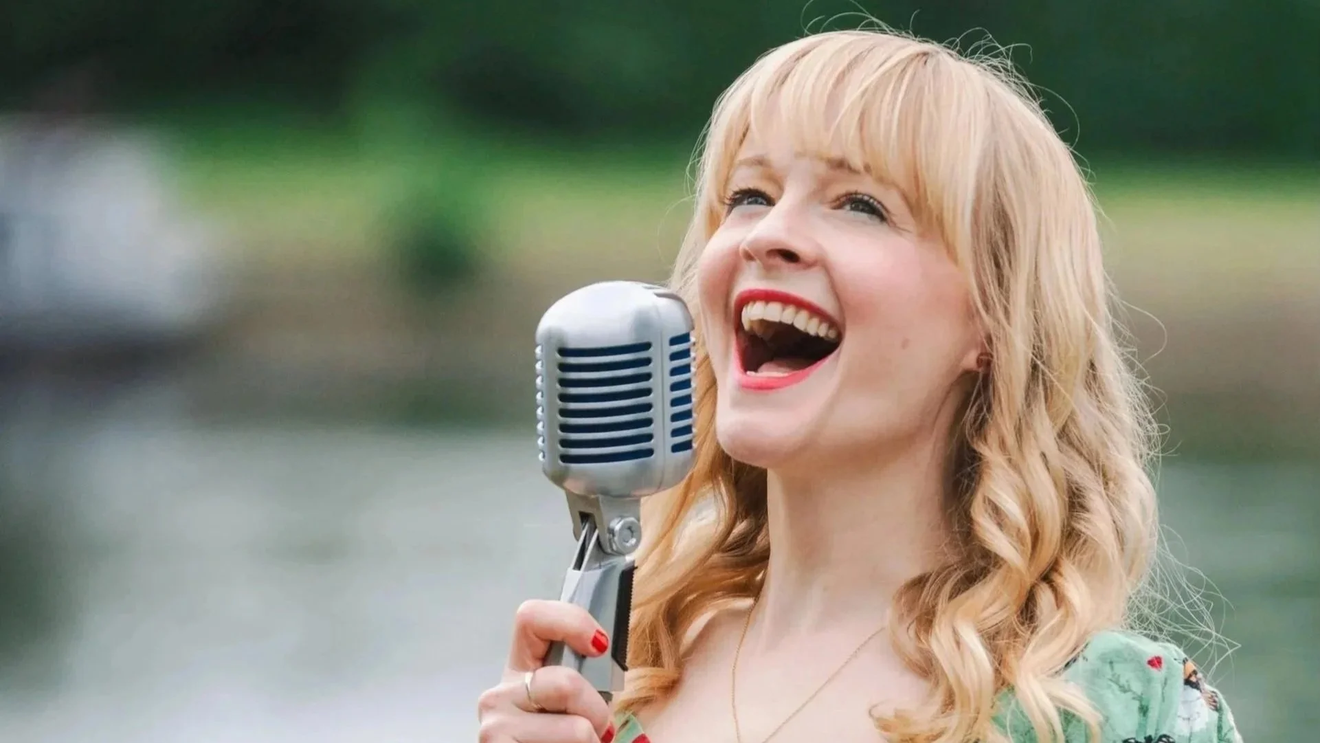 Kitty Mazinsky sings, smiling, into a stylish vintage microphone (a Shure 55). In the background is the river Thames. Kitty wears her long blonde hair in loose curls, with the shoulders of a green dress just visible, and her nails painted scarlet..