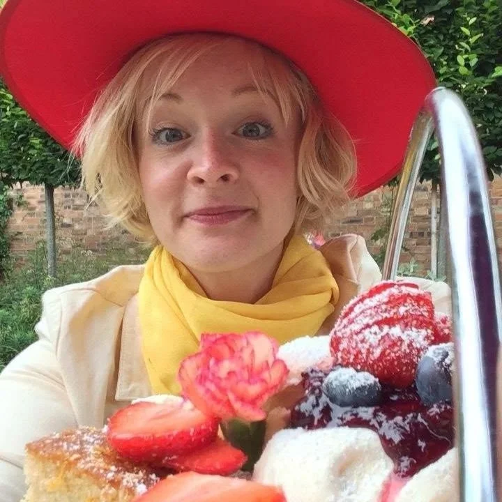 Kitty Mazinsky wearing a wide-brimmed red hat and yellow scarf, photographed in the garden at the Folly Tea Rooms, Holt, Norfolk, with an afternoon tea stand in the foreground