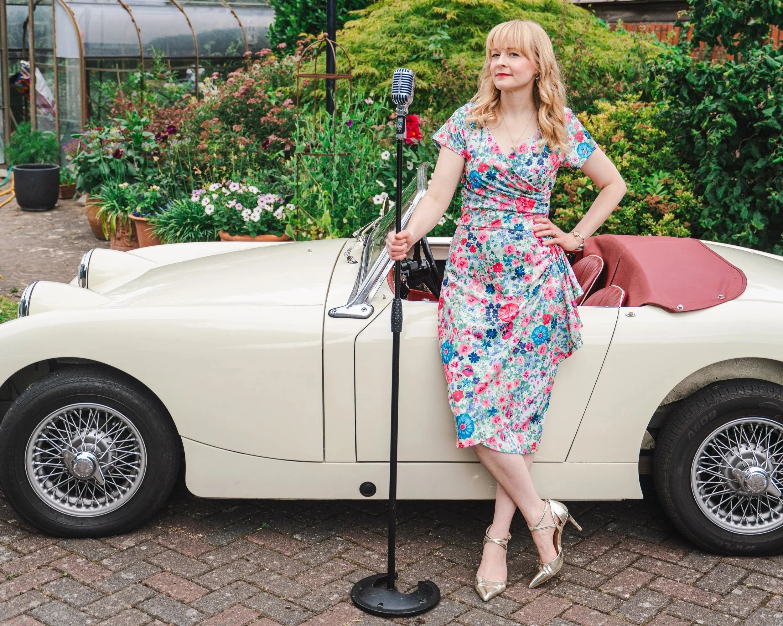 Kitty Mazinsky leans on an Austin Healey frogeye sprite in a lush garden setting with much greenery, potted plants, and a greenhouse.. She holds a mic stand topped with a stylish vintage Shure 55 mic and wears a fitted knee-length floral dress..