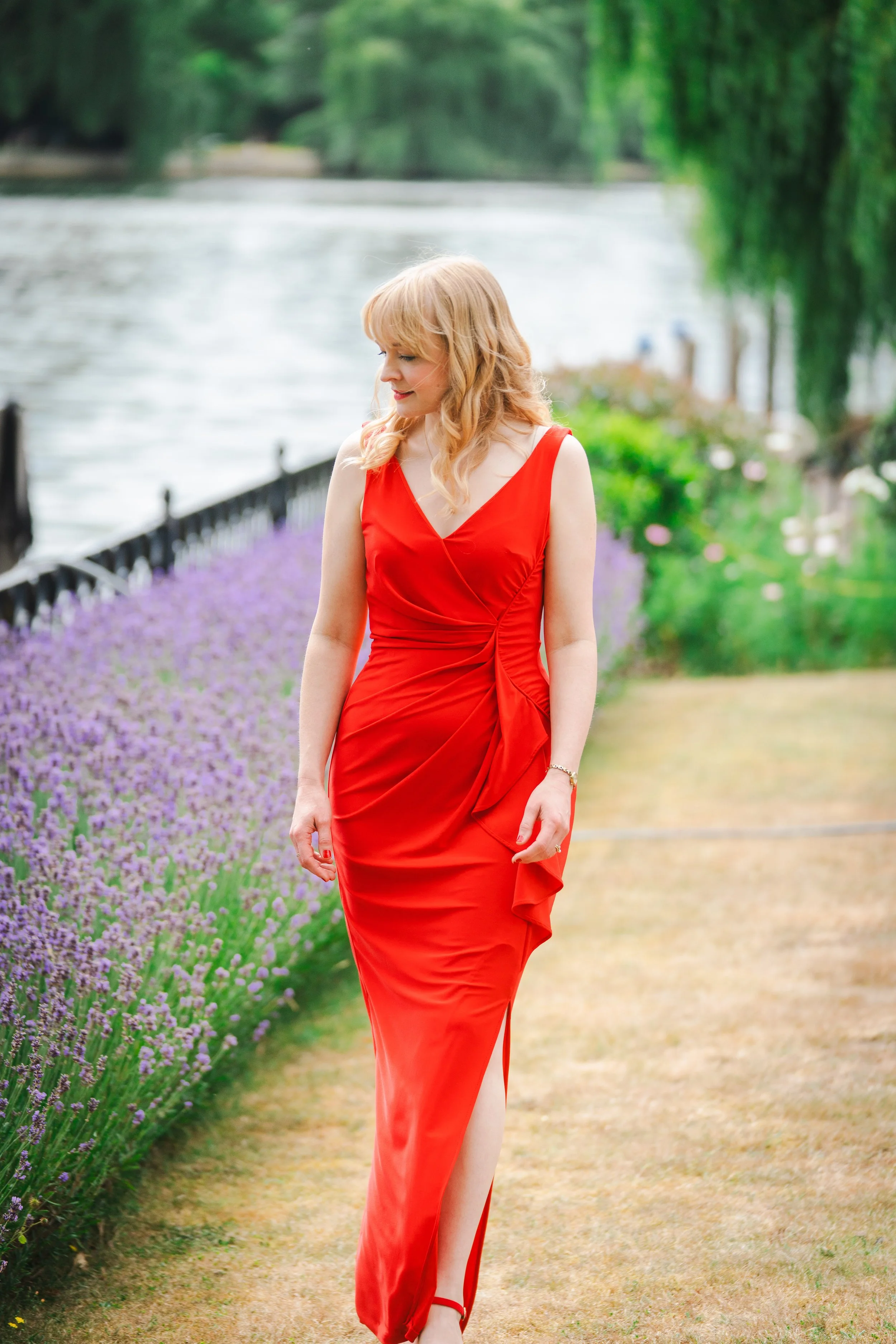 Kitty Mazinsky wears a scarlet evening dress, walking alongside a bed of lavender in a garden setting alongside a river.