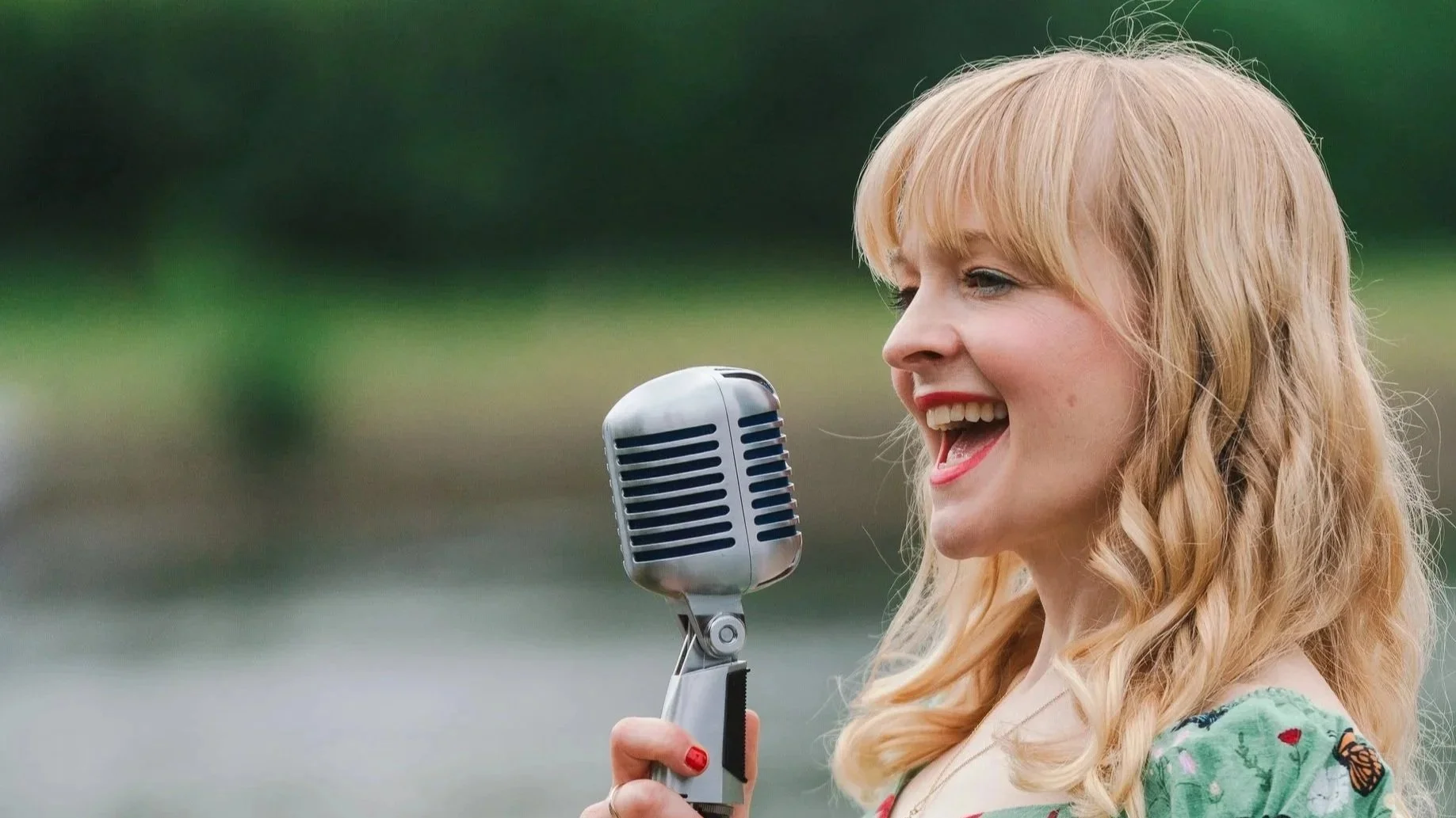 Kitty Mazinsky sings into a stylish vintage microphone, her blonde hair falling in loose curls. In the background is the river Thames.