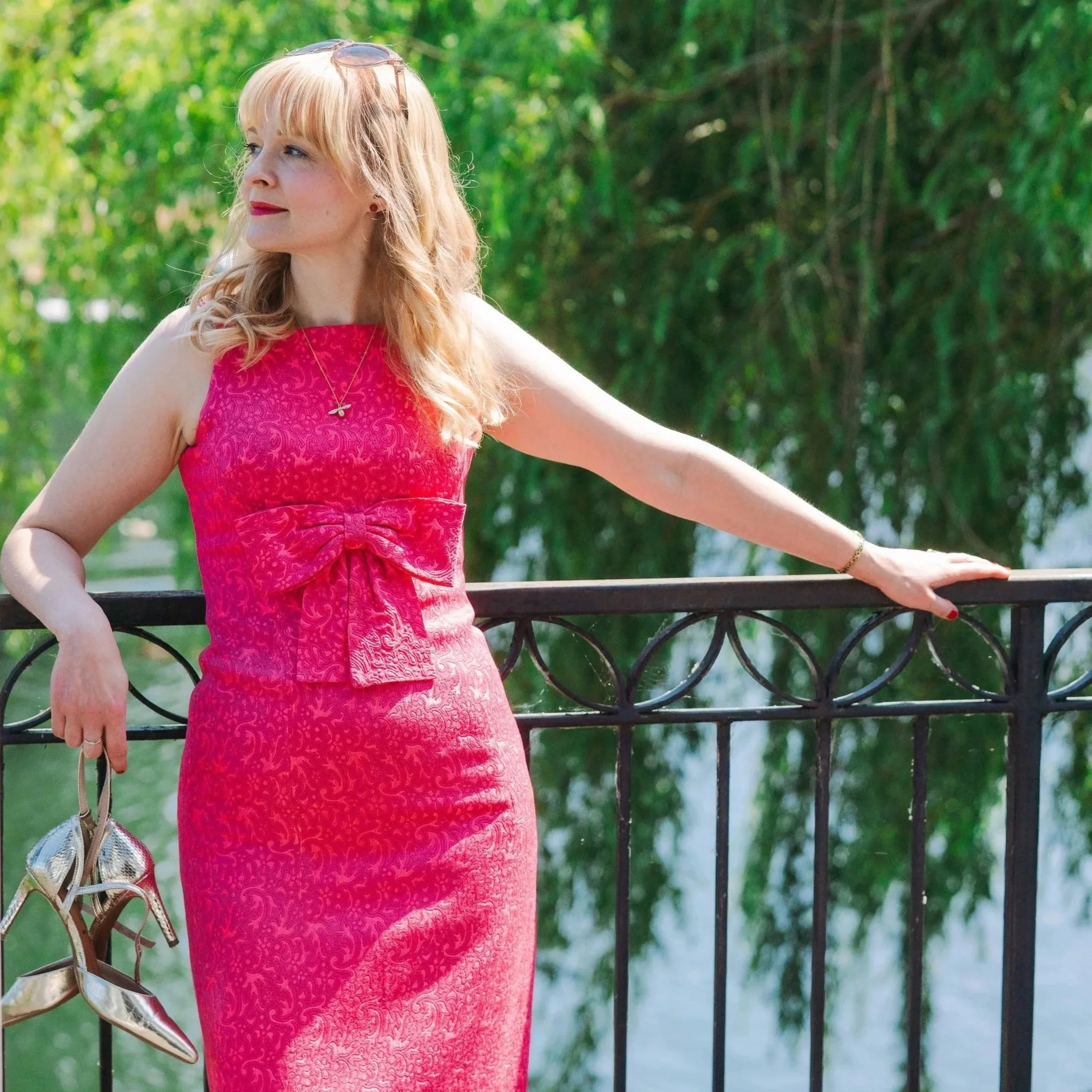 Kitty Mazinsky wears a flamingo-pink, floor-length 1960s sleeveless evening dress with a bow at the waist, standing on a balcony in front of a weeping willow that gently reaches down over the Thames