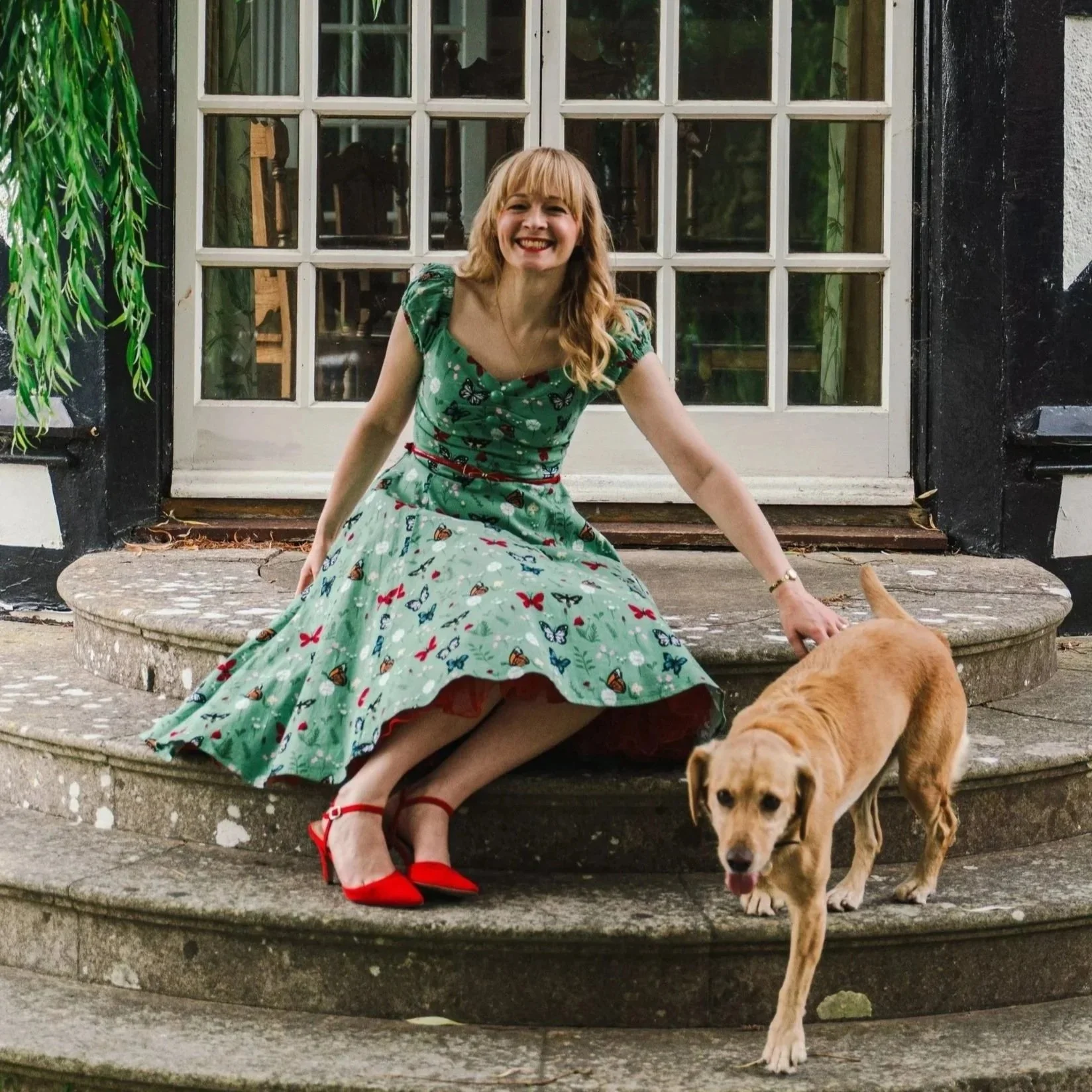 Kitty Mazinsky sits on the crescent-shaped stone steps of a Berkshire country house, a weeping willow to the left of the frame. She has long blonde hair falling in open curls, and is reaching out to touch a Labrador descending the steps.