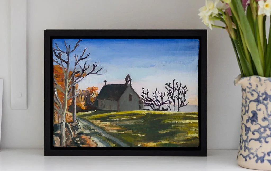Painting of a rural landscape with a small church, leafless trees, a grassy field, and a partly cloudy sky, framed and displayed on a white surface next to a blue and white patterned vase with daffodils.