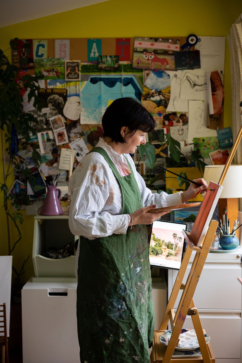 Woman painting on canvas in art studio with collage of artworks and photographs on wall behind her.