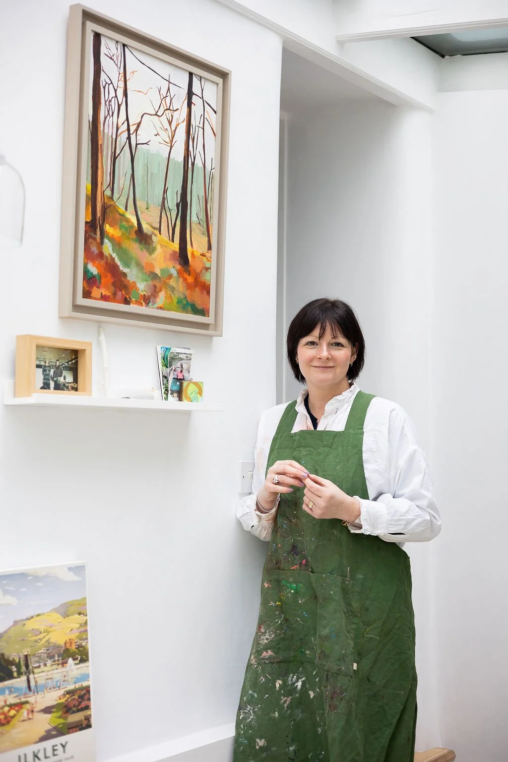 Woman with short dark hair wearing a white shirt and green paint-stained apron standing in an art gallery, smiling at the camera, with a colorful landscape painting of trees hanging on the white wall behind her.