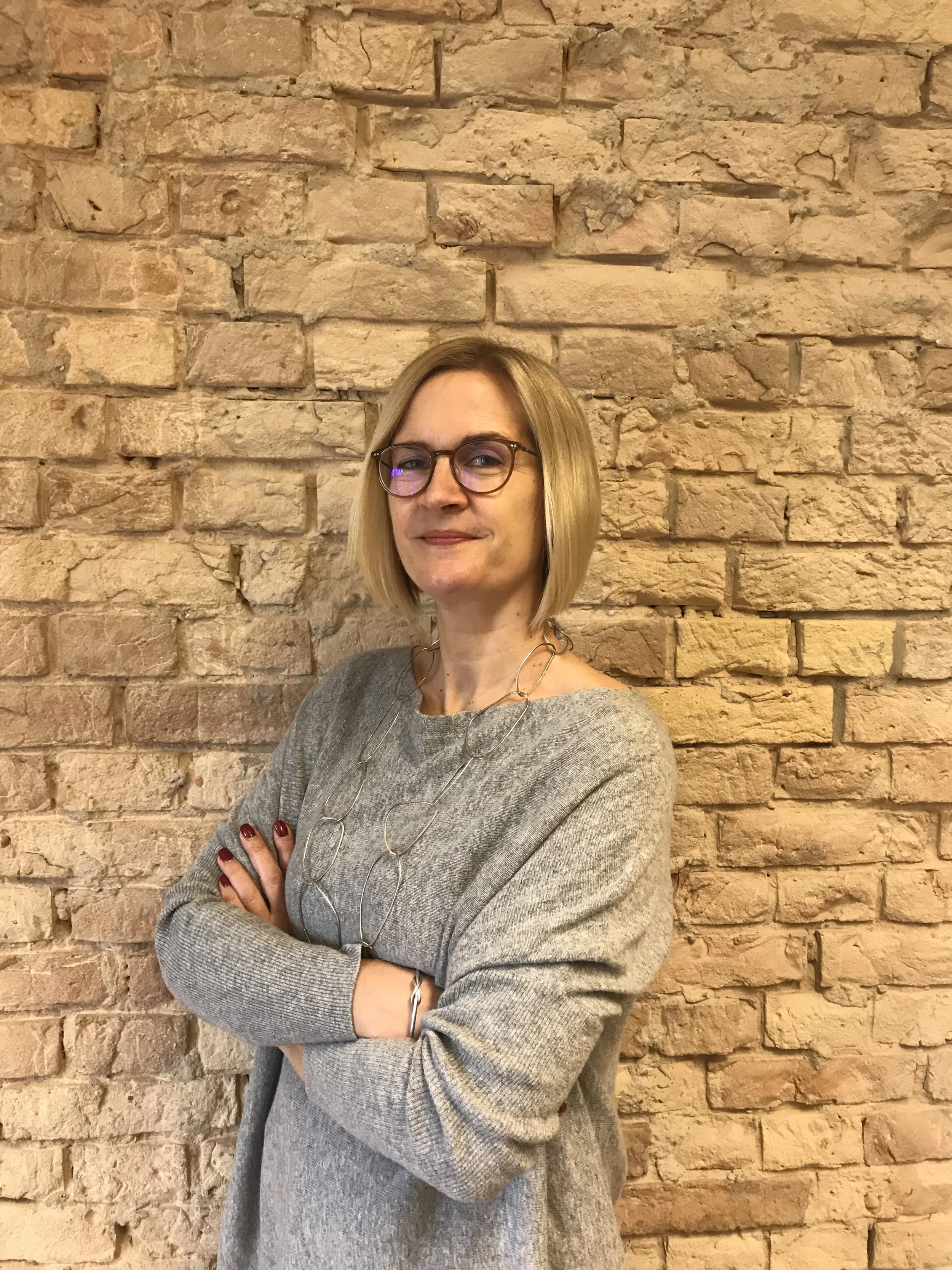 Eine Frau mit Brille und blonden Haaren steht vor einer Ziegelmauer, trägt einen grauen Pullover und eine silberne Kette.