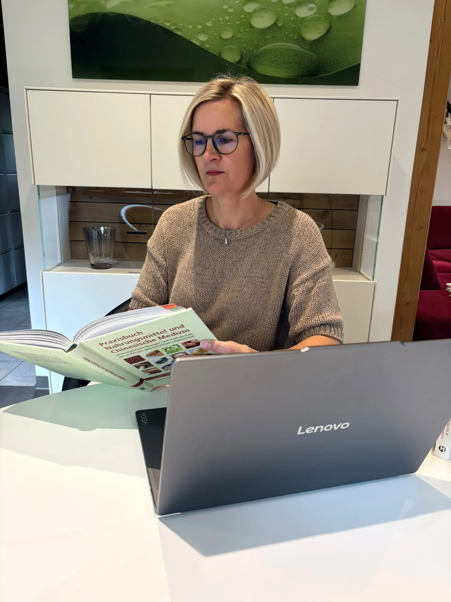 Eine Frau mit Kurzhaarschnitt und Brille liest ein Gesundheits- und Ernährungshandbuch, während sie an einem weißen Tisch mit einem aufgeschlagenen Laptop sitzt.