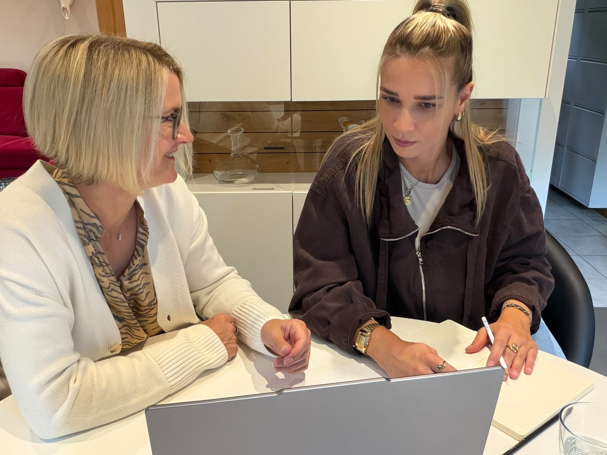Zwei Frauen sitzen an einem Tisch, eine ältere Frau mit Brille und blonde Haare im weißen Pullover, eine jüngere Frau mit langen blonden Haaren in einem braunen Jacke, schreiben auf Papier und diskutieren.