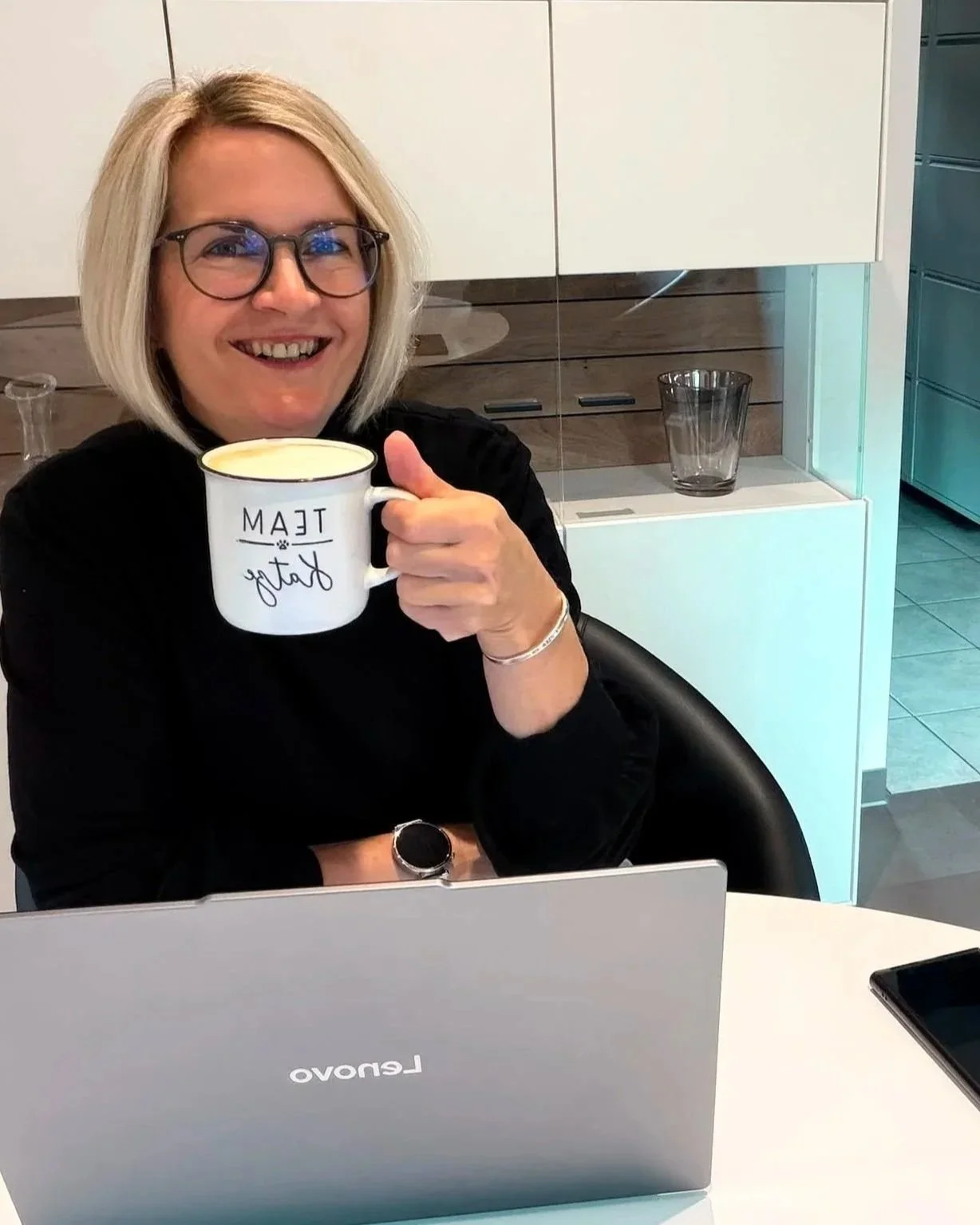 Frau mit Brille und einem Lächeln hält eine weiße Tasse mit der Aufschrift 'Team' in einer modernen Küche.