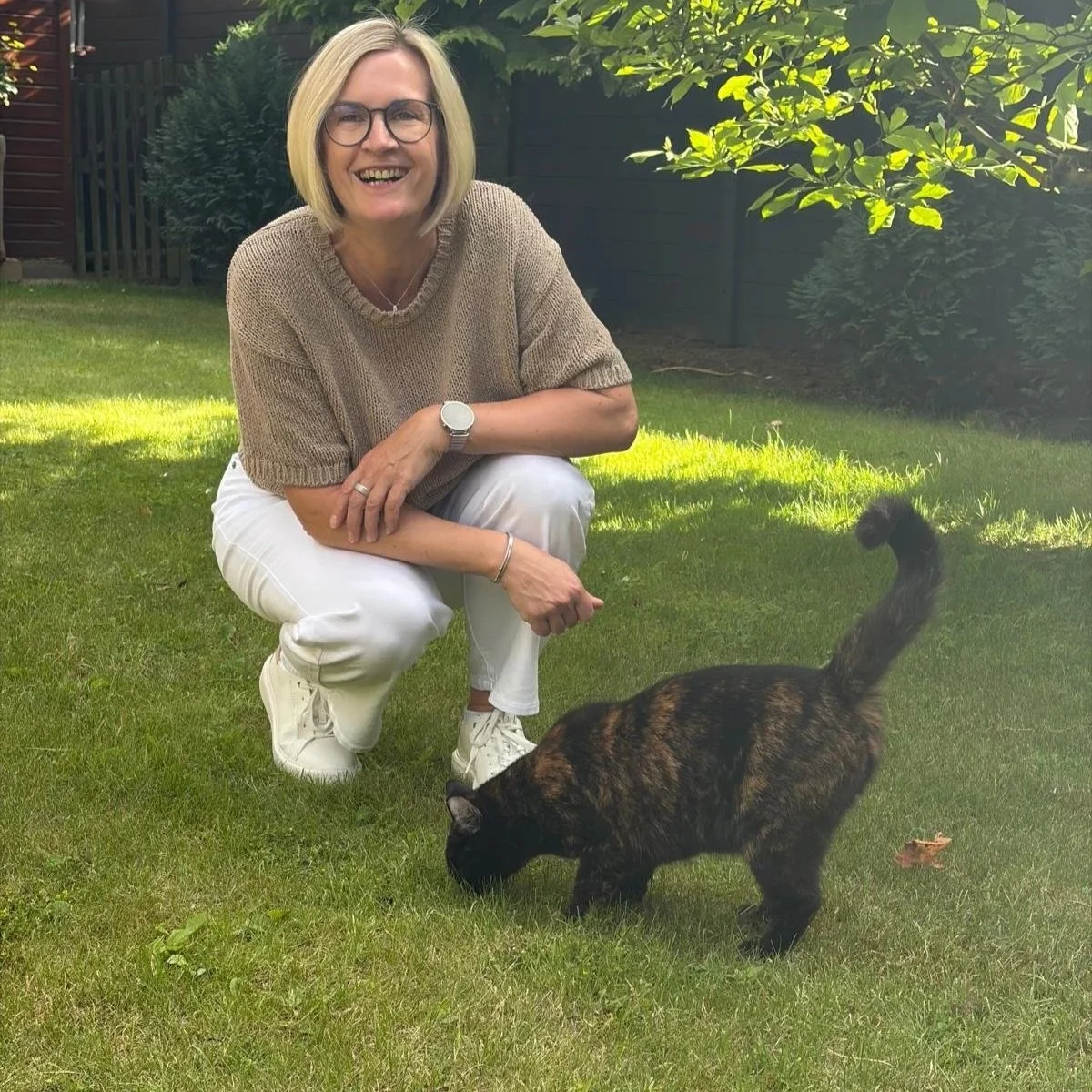 Eine lachende Frau mit Brille und kurzen blonden Haaren kniet auf dem Gras im Garten, während eine schwarze und braune gestreifte Katze also Schnurrt; im Hintergrund sind Bäume und ein Zaun zu sehen.