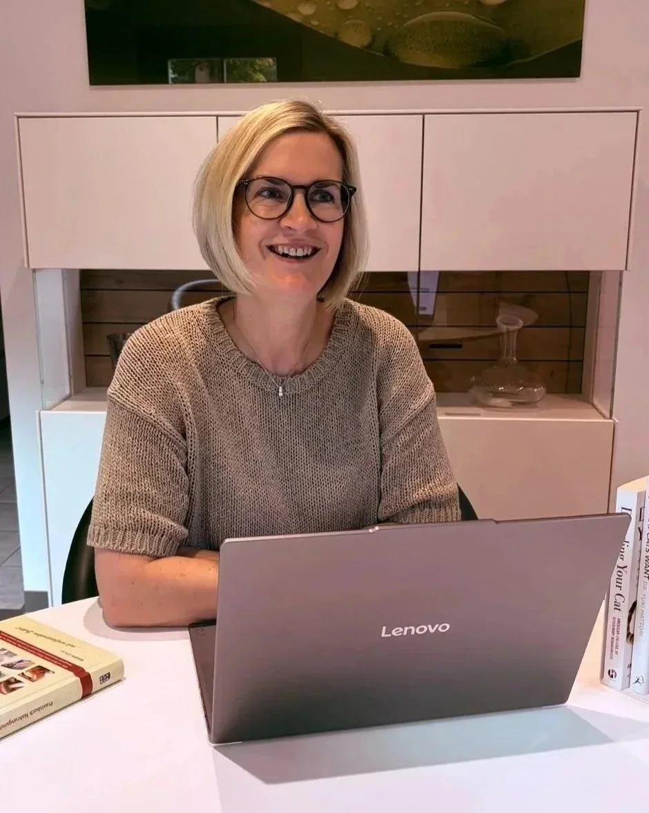 Eine lachende Frau mit Brille sitzt an einem Tisch mit einem offenen Lenovo-Laptop, neben ihr liegen Bücher.