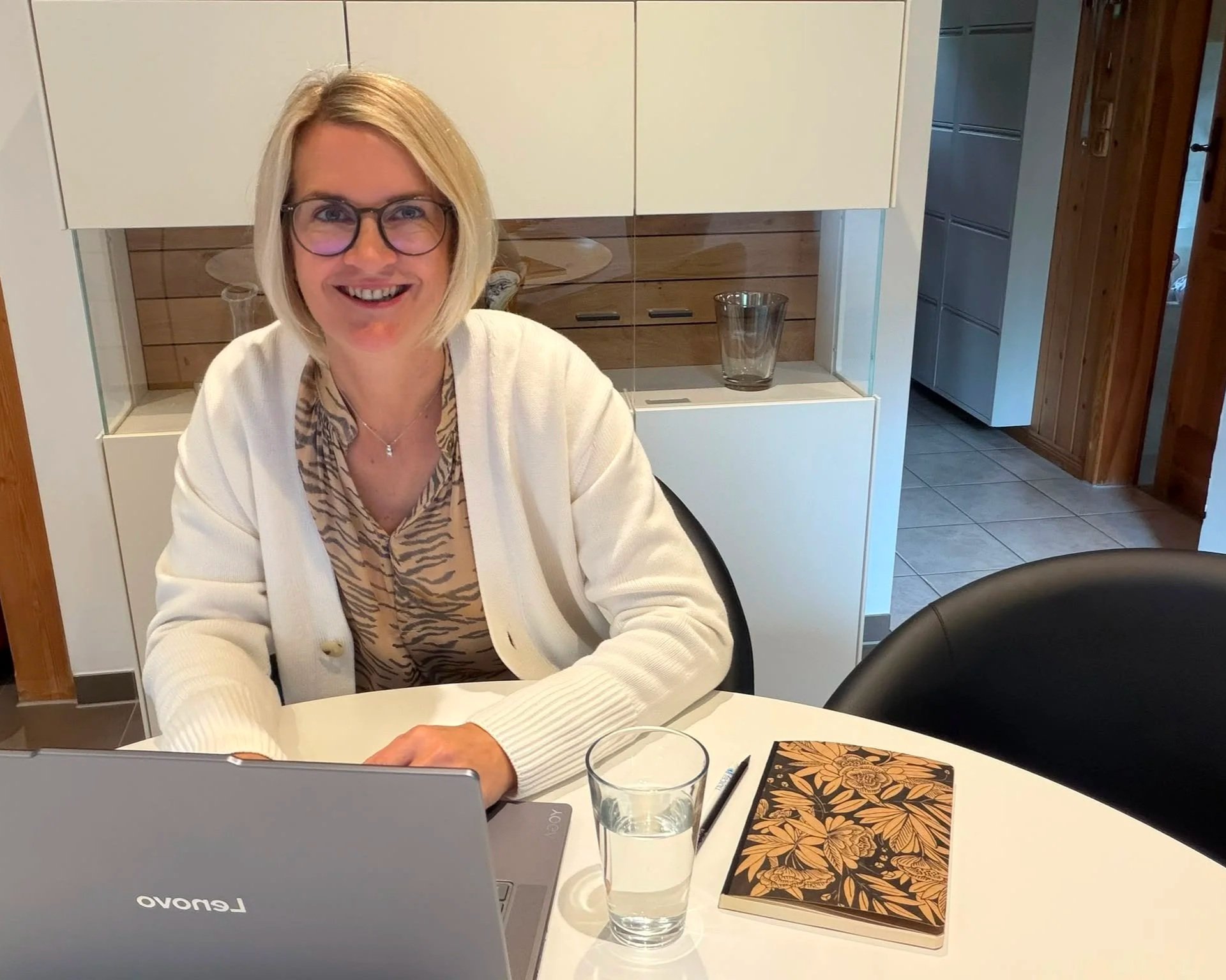 Eine lachende blonde Frau mit Brille sitzt an einem Esstisch mit einem Laptop, einem Glas Wasser, einem Buch mit floralem Muster und einem Stift.