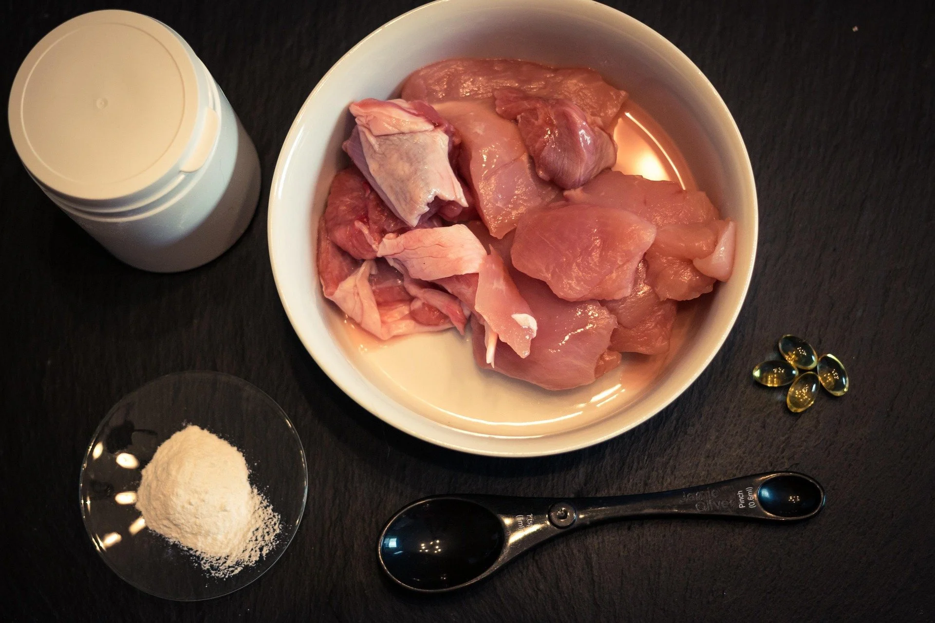 Schüssel mit rohem, gewürfeltem Fleisch, Flasche mit Gelatine, kleine Schale mit Gewürzpulver, Löffel, schwarze Kapseln, und Messlöffel auf dunklem Tisch.