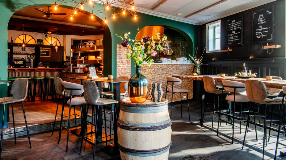 Interieur van een restaurant met een bar en hoge tafels, versierd met bloemvasen en sfeervol licht.
