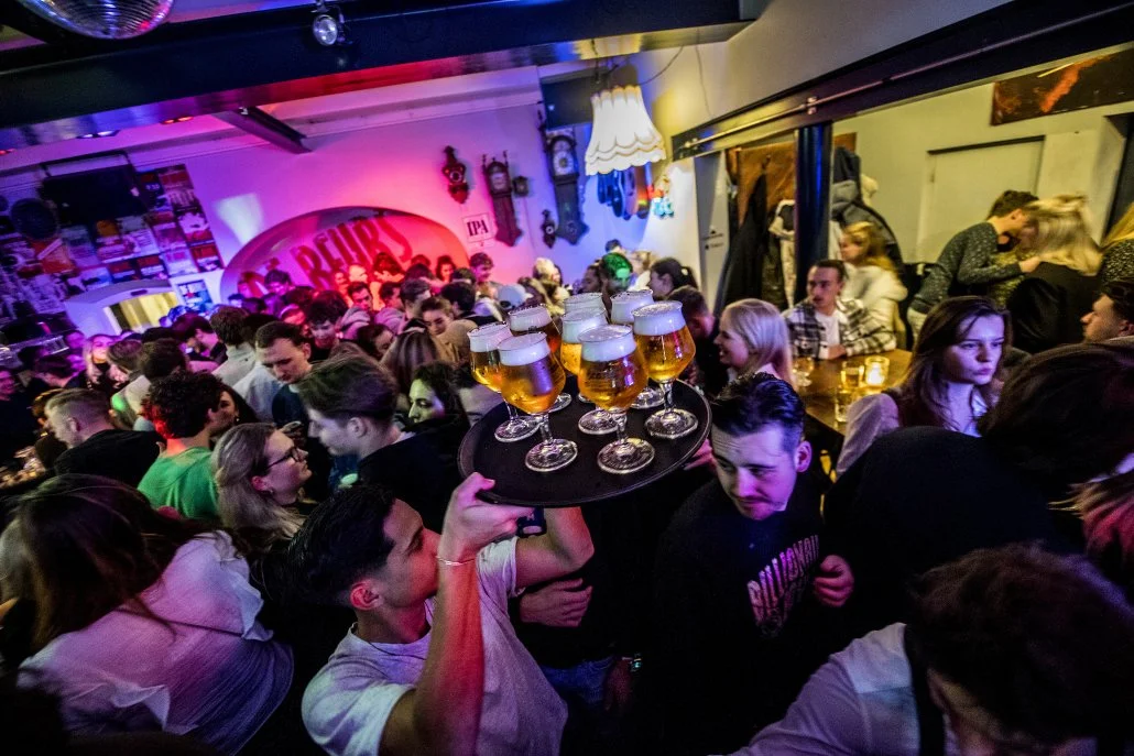 Een drukke bar of club met veel mensen die genieten en socializen, een dienblad met acht glazen bier wordt door een ober door de menigte gedragen.