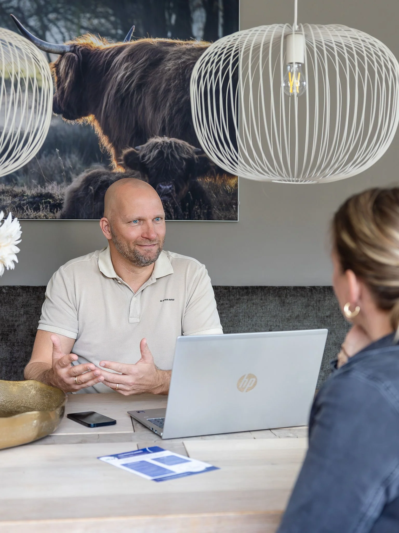 Twee mensen in gesprek aan een tafel met een laptop, een man en een vrouw, met een posters van bizons op de muur en lichtarmaturen erboven.