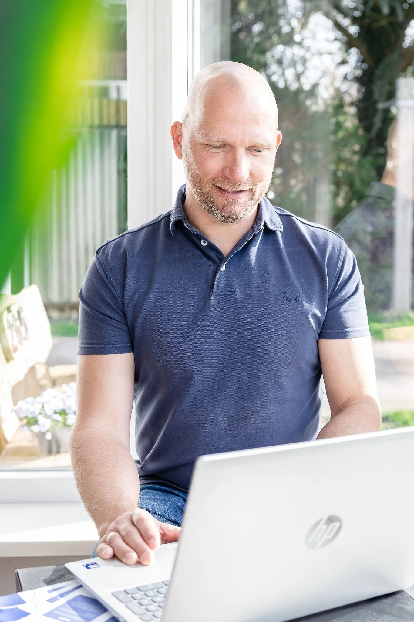 Man zit bij een raam en kijkt naar een laptop, met spiegels in de achtergrond en zonlicht dat binnenkomt.
