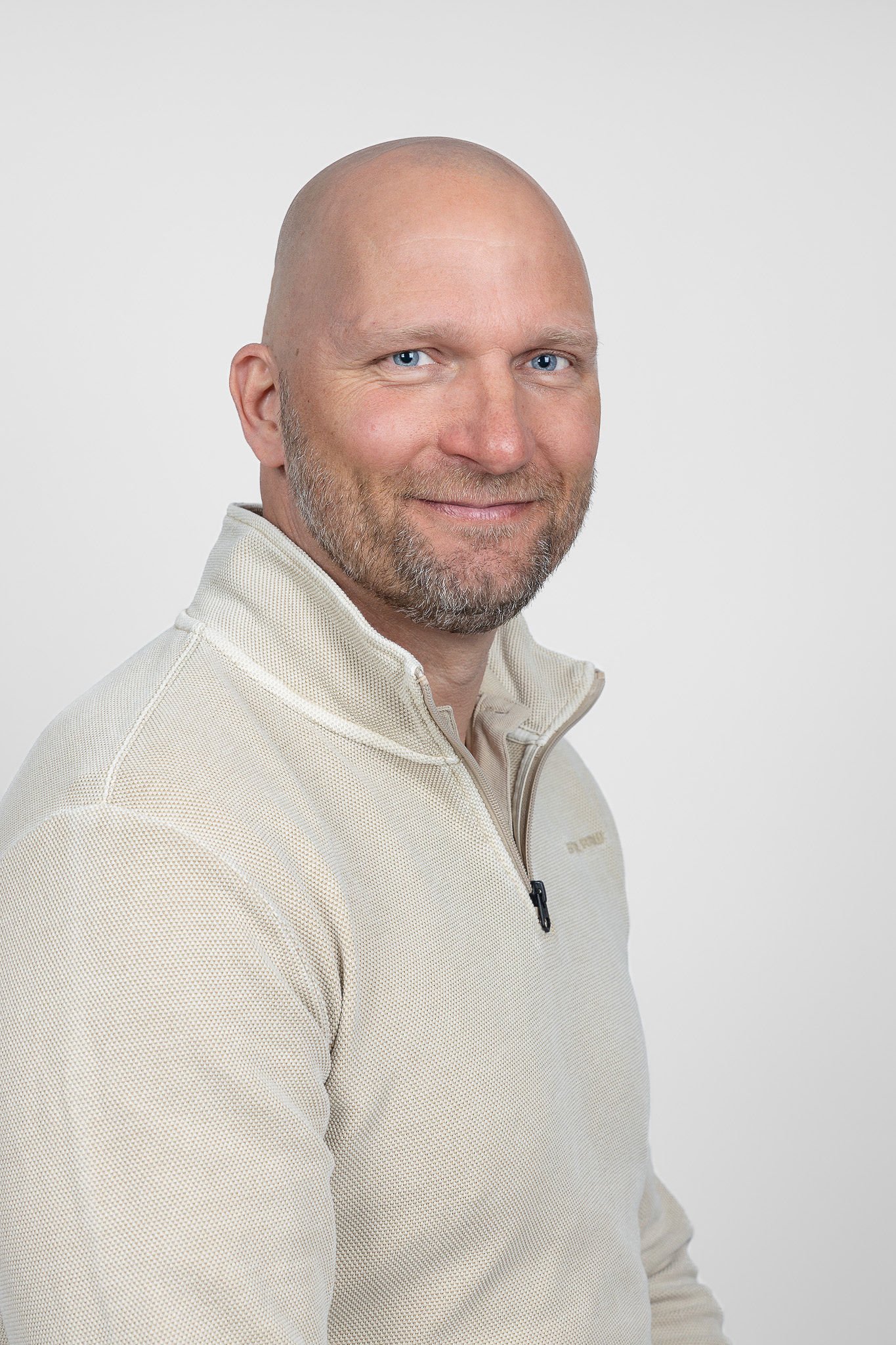 Portretfoto van een man met een baard en een vriendelijke glimlach, gekleed in een beige trui met rits, tegen een witte achtergrond.
