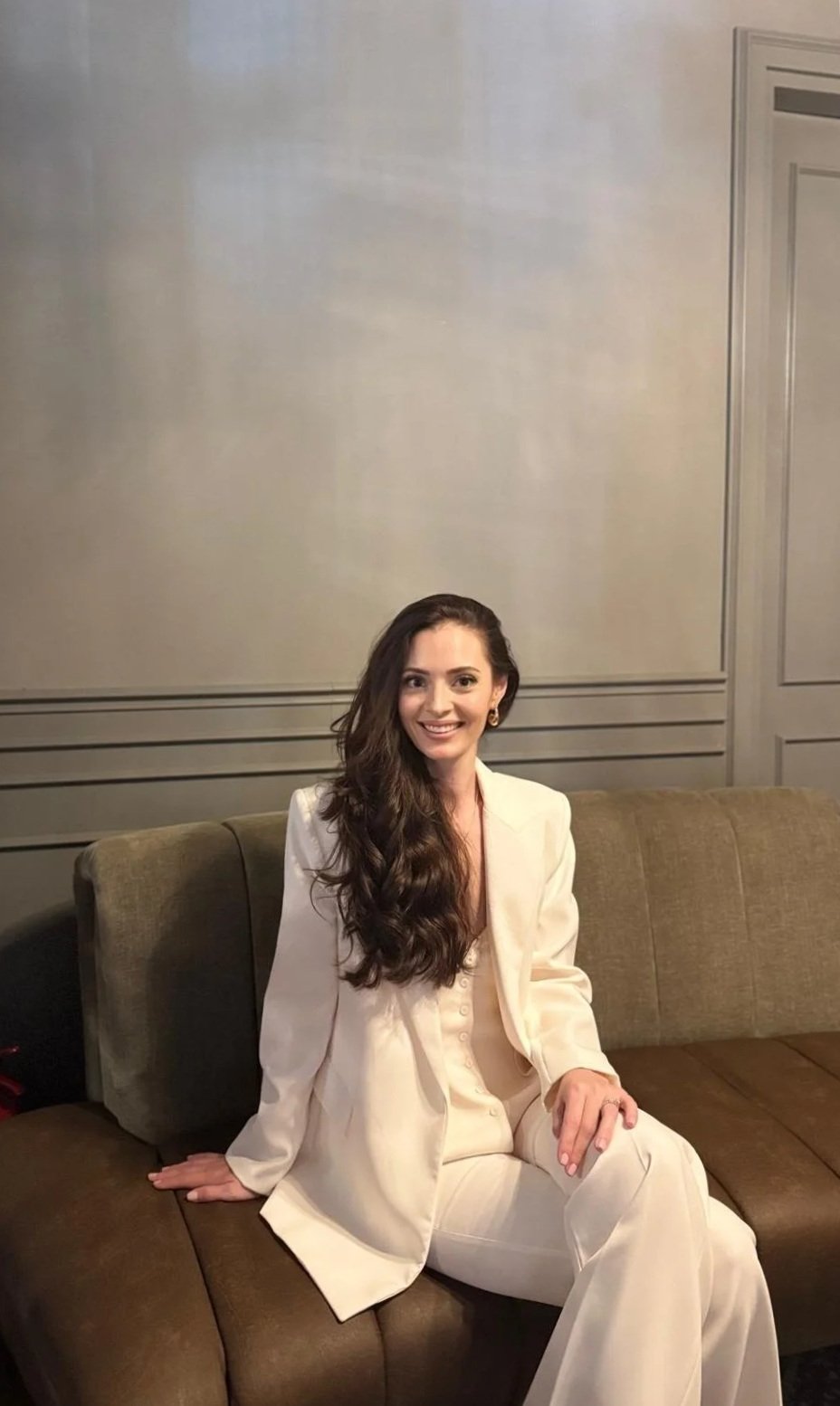Woman with long brown hair wearing a cream-colored suit sitting on a brown sofa, smiling at the camera, in a room with gray paneled walls.