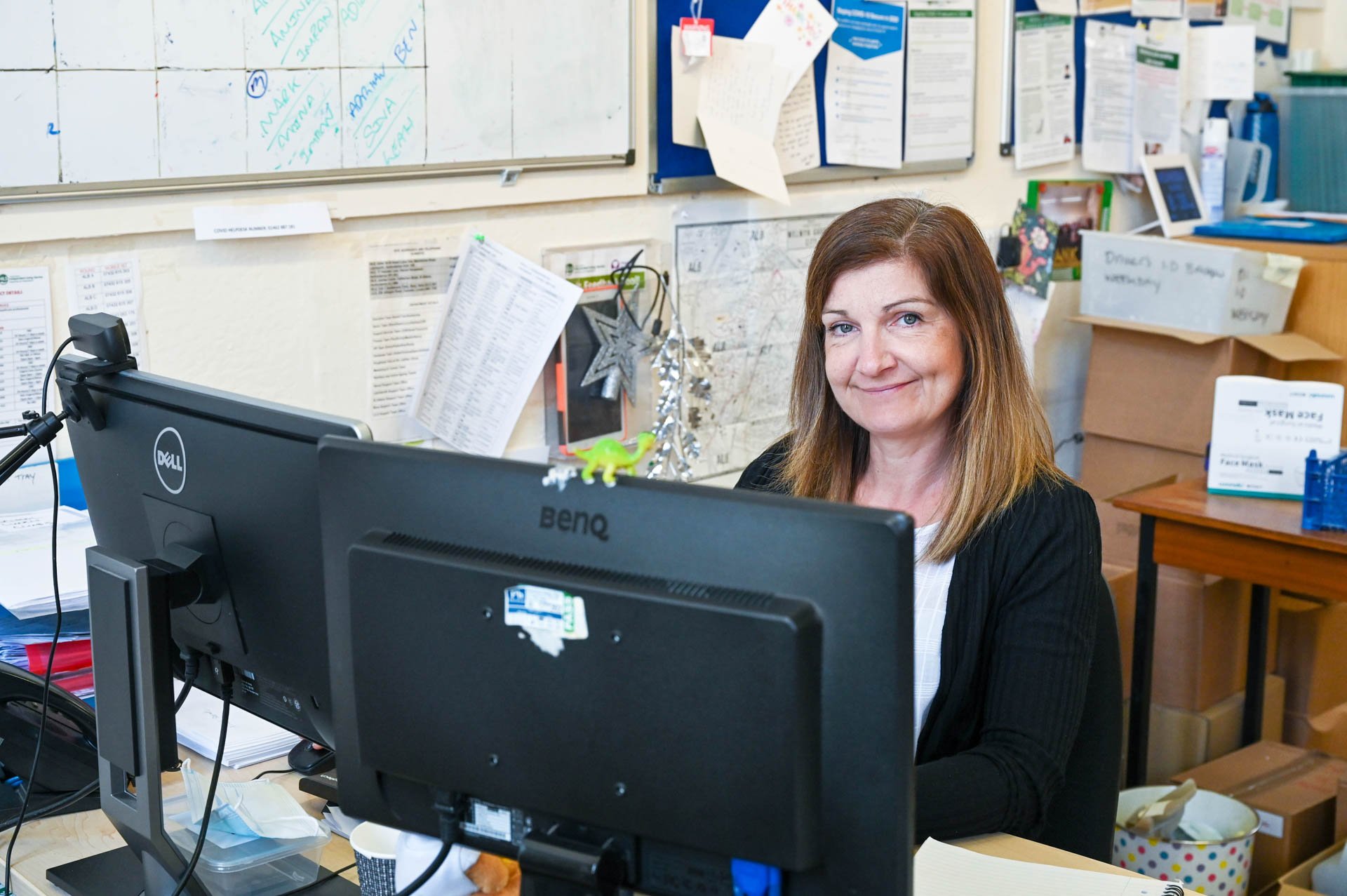 A woman with shoulder-length brown hair sitting at a cluttered office desk with dual computer monitors, smiling at the camera.
