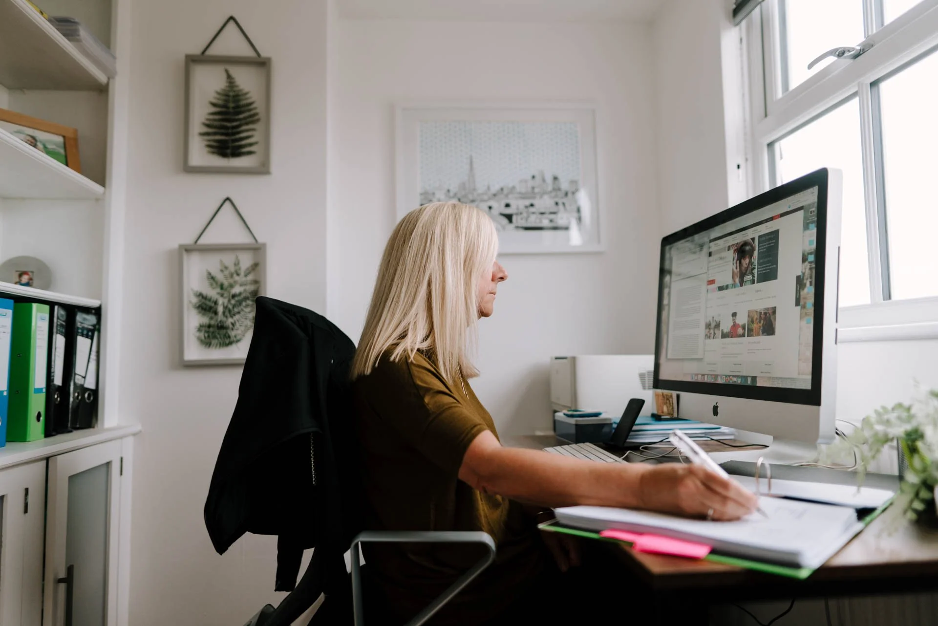 A woman working at her desk in a home office, using a computer and taking notes with a pen. There are bookshelves, framed botanical pictures, and a window in the background.