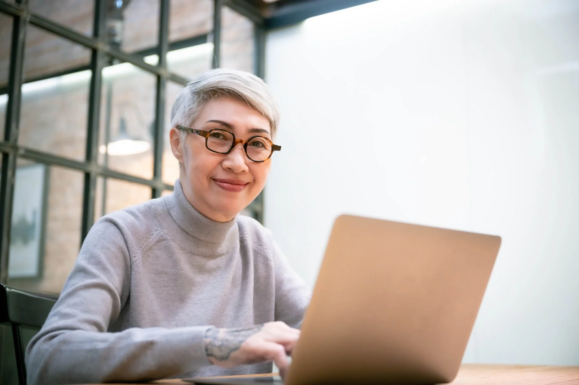 Smiling woman with short gray hair and glasses working on a laptop in a modern office with large windows.