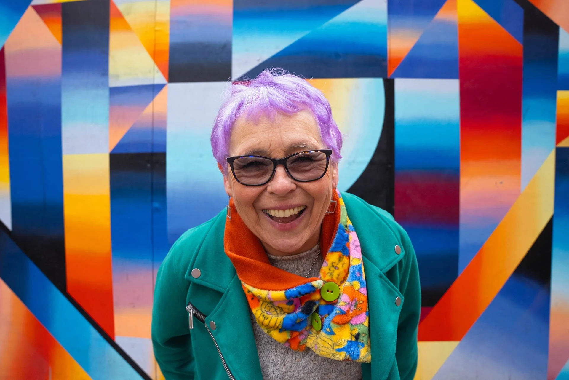 Older middle aged woman with short purple hair, laughing at the camera, wearing a brightly coloured scarf, green leather jacket and standing in front of a multi-coloured background