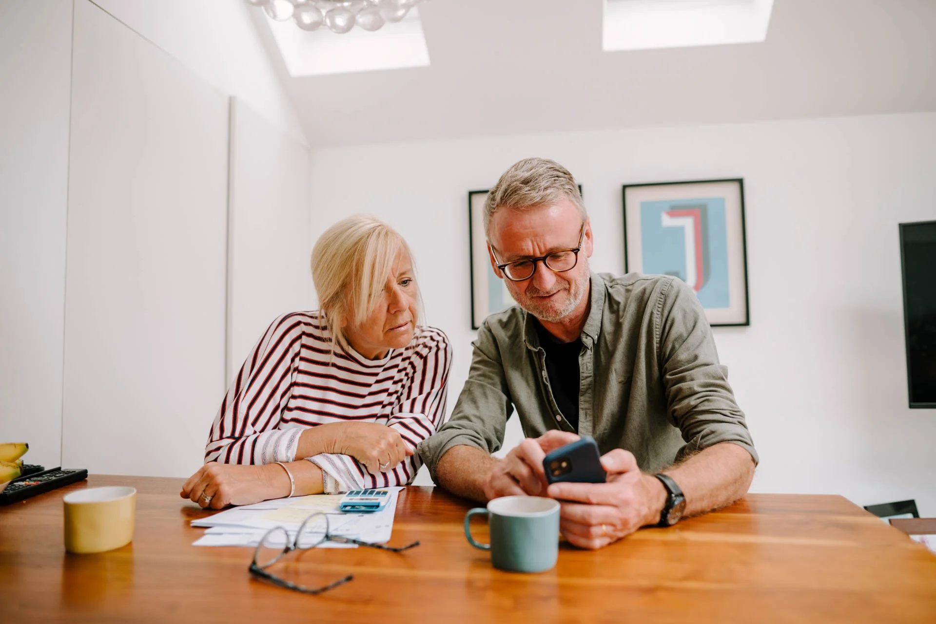 An older woman and man sitting at a wooden table in a bright room, looking at a smartphone the man is holding. The woman appears curious, and the man is smiling. On the table are papers, a calculator, glasses, a cup, and a banana. Behind them, framed abstract artwork hangs on a white wall.