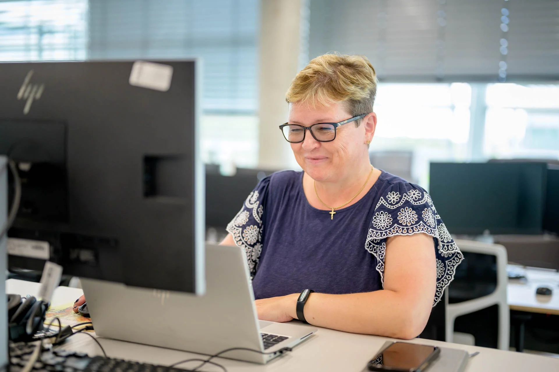 Woman working at her desk in an office, using a laptop and looking at a desktop monitor, with bright windows in the background.