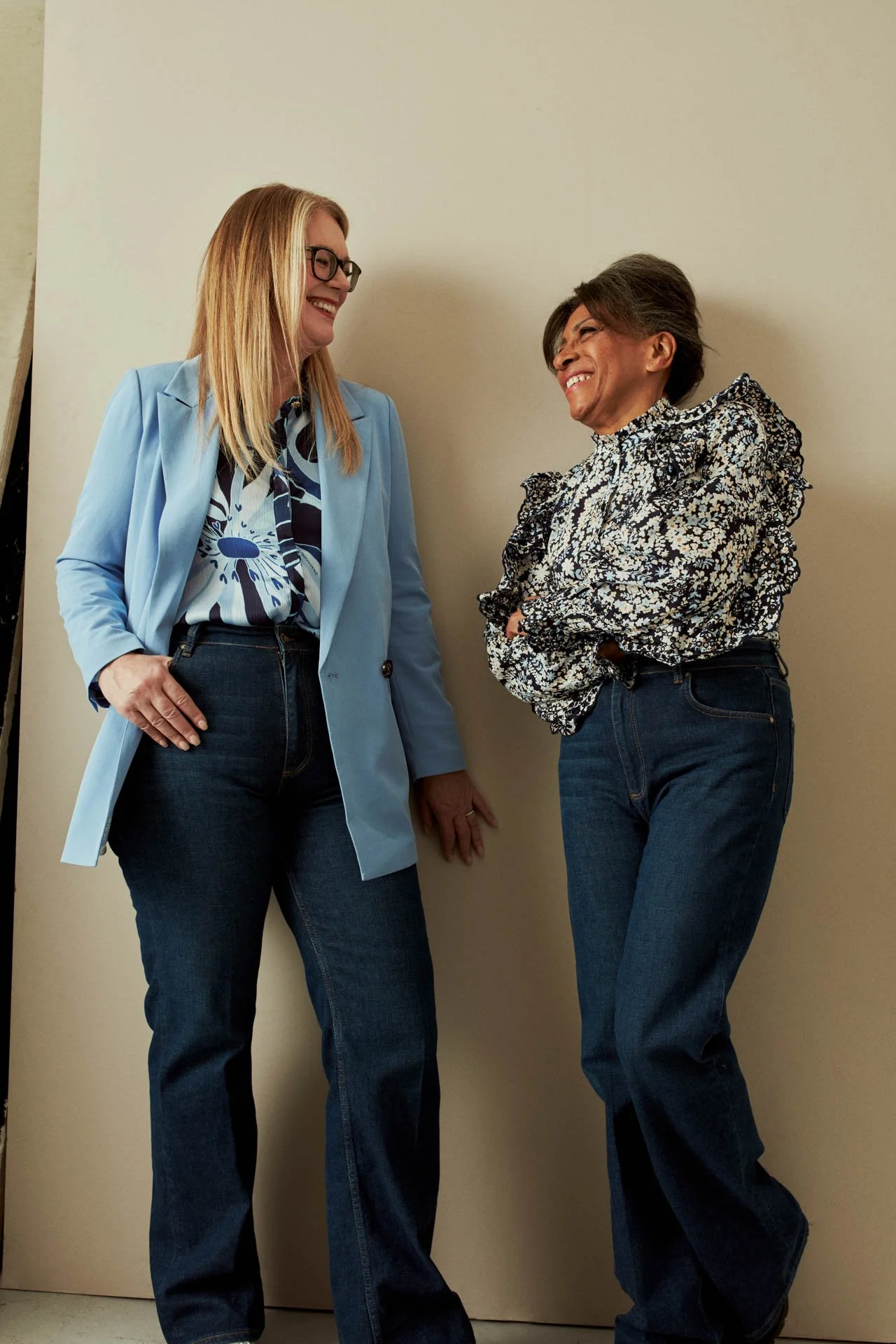 Two women are standing and talking with each other, smiling. One is wearing a light blue blazer and glasses, the other is wearing a patterned blouse with ruffles and jeans. They are indoors against a plain beige wall.